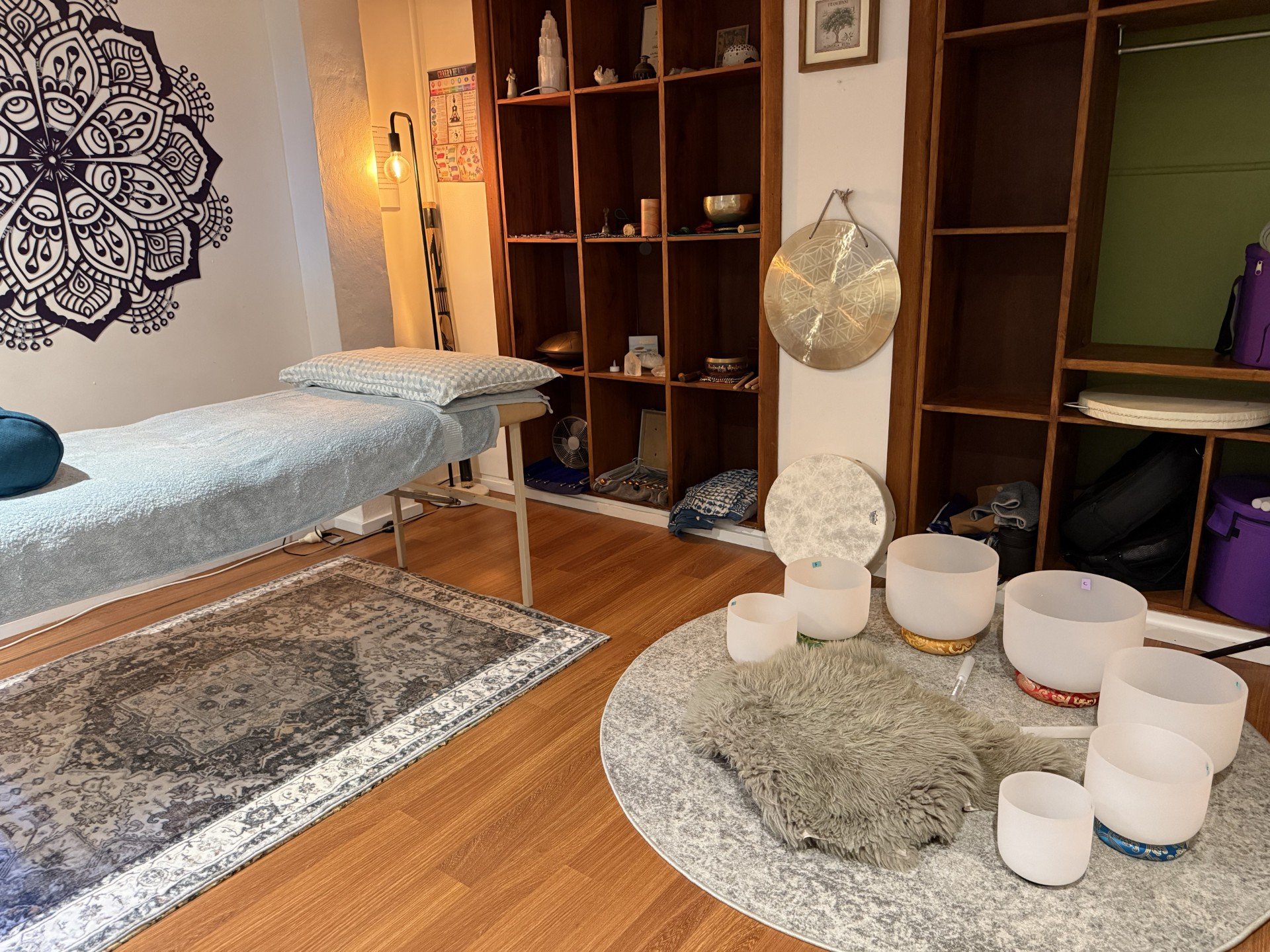 Sound Stone Therapy healing space. Room with wooden flooring, a massage table with a pillow, a patterned rug, a shelving unit with decorations, a large gong, singing bowls, and musical instruments, near a white wall with a black decorative wall art.