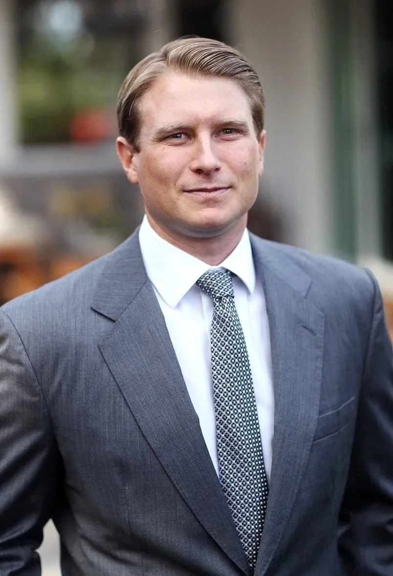 A young man in a gray business suit with a patterned tie, standing outdoors, smiling slightly.