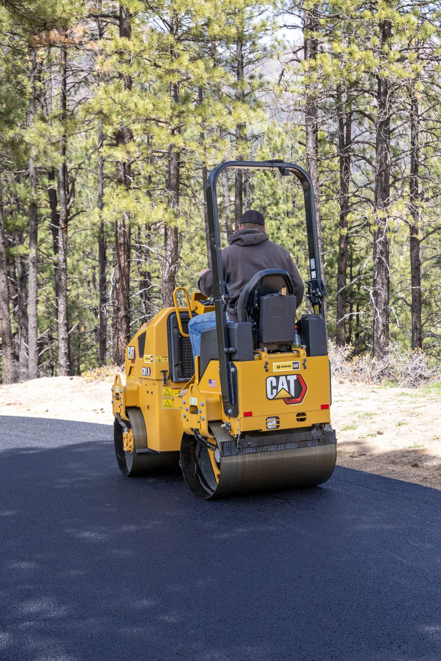 Man operating a yellow Caterpillar road roller compacting freshly paved black asphalt on a sunny day, surrounded by a forest of pine trees.