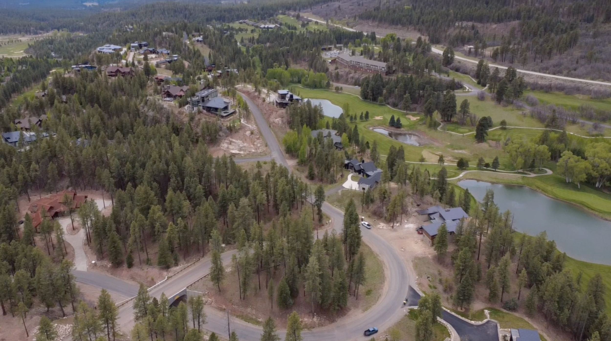 Aerial view of a residential area surrounded by trees, with winding roads, a pond, and houses with dark roofs.