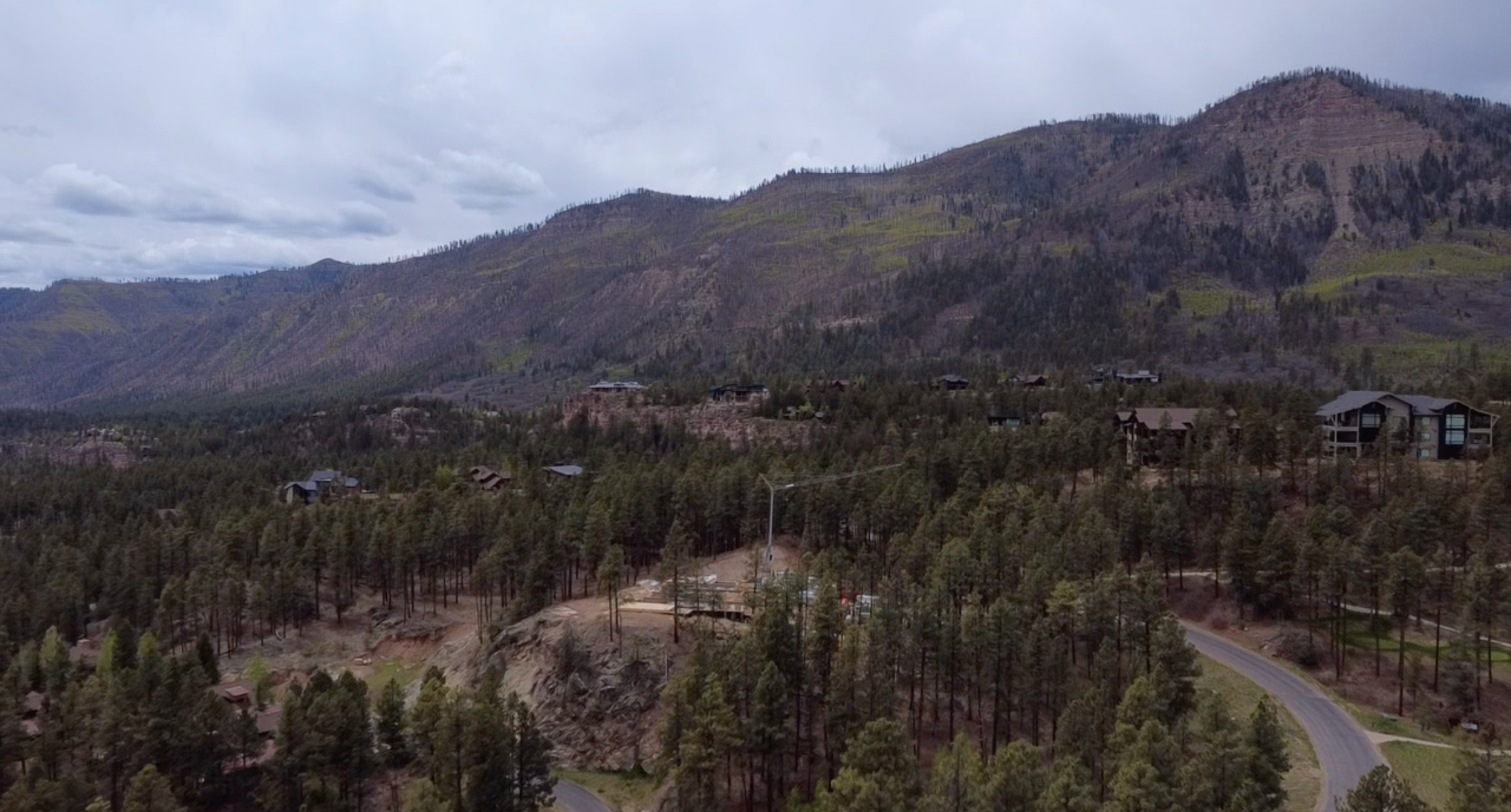 Mountain landscape with scattered houses, trees, and winding dirt and paved roads under a cloudy sky.