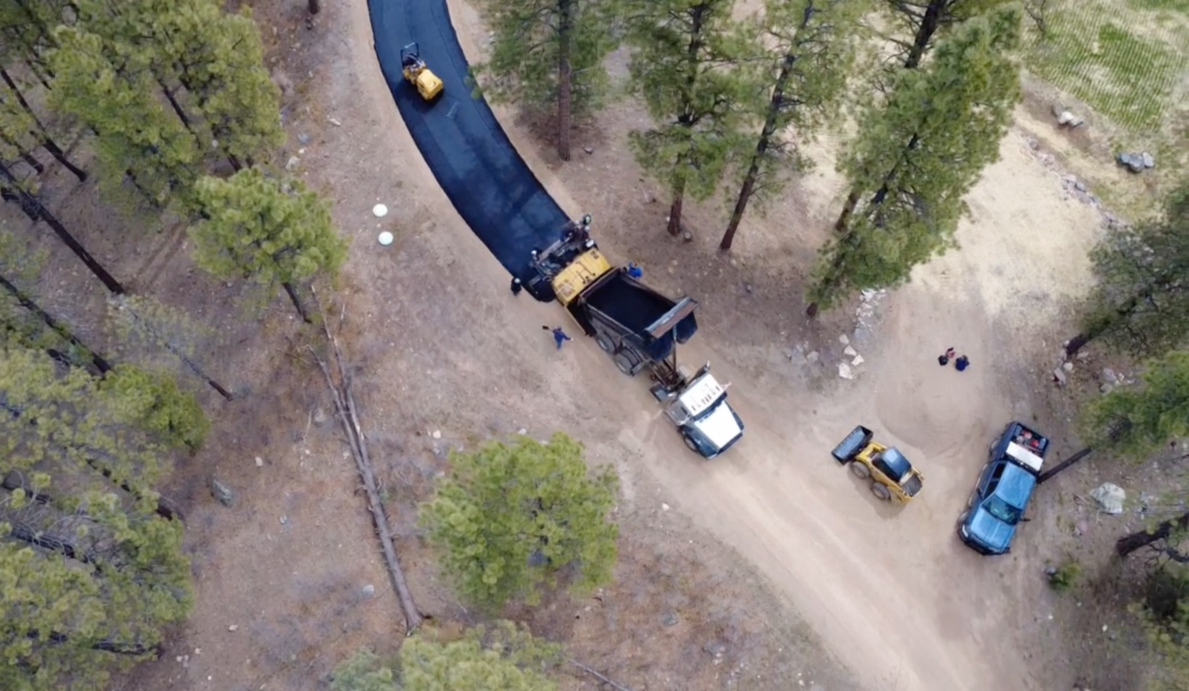An aerial view of a forested area with a dirt road or pathway. There are several vehicles and construction equipment along the road, including a road roller and a dump truck. Trees surround the area, and there are some rocks and scattered objects on 