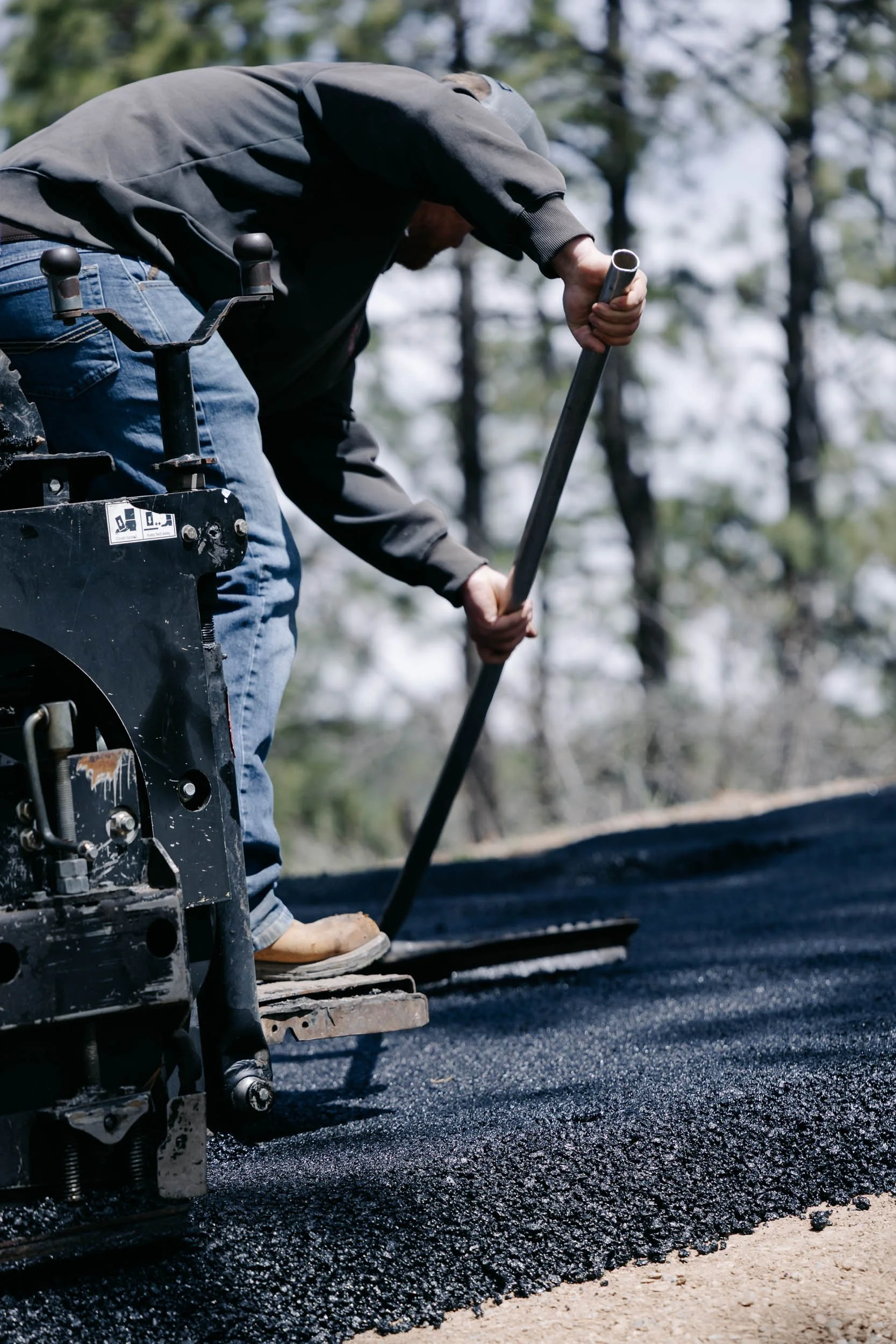 Worker with a gray hoodie and jeans operating a machine to lay fresh asphalt on a road during daytime, with trees in the background.