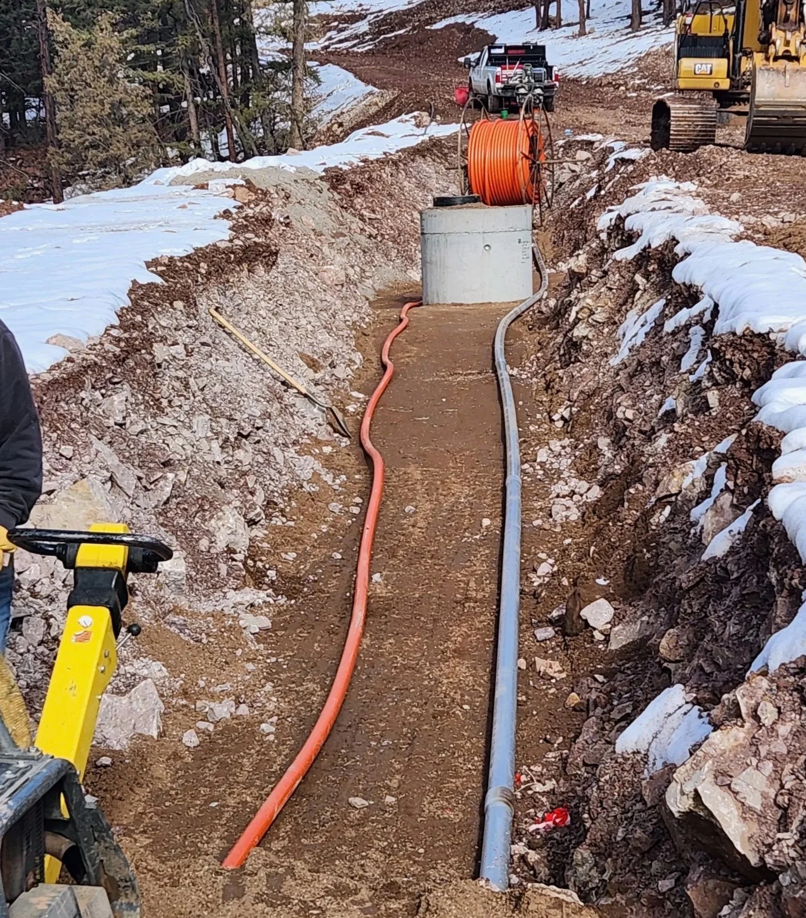 Construction site with dirt road, orange hose reel on a concrete block, yellow piece of equipment in the foreground, and trucks in the background, snow on the ground.