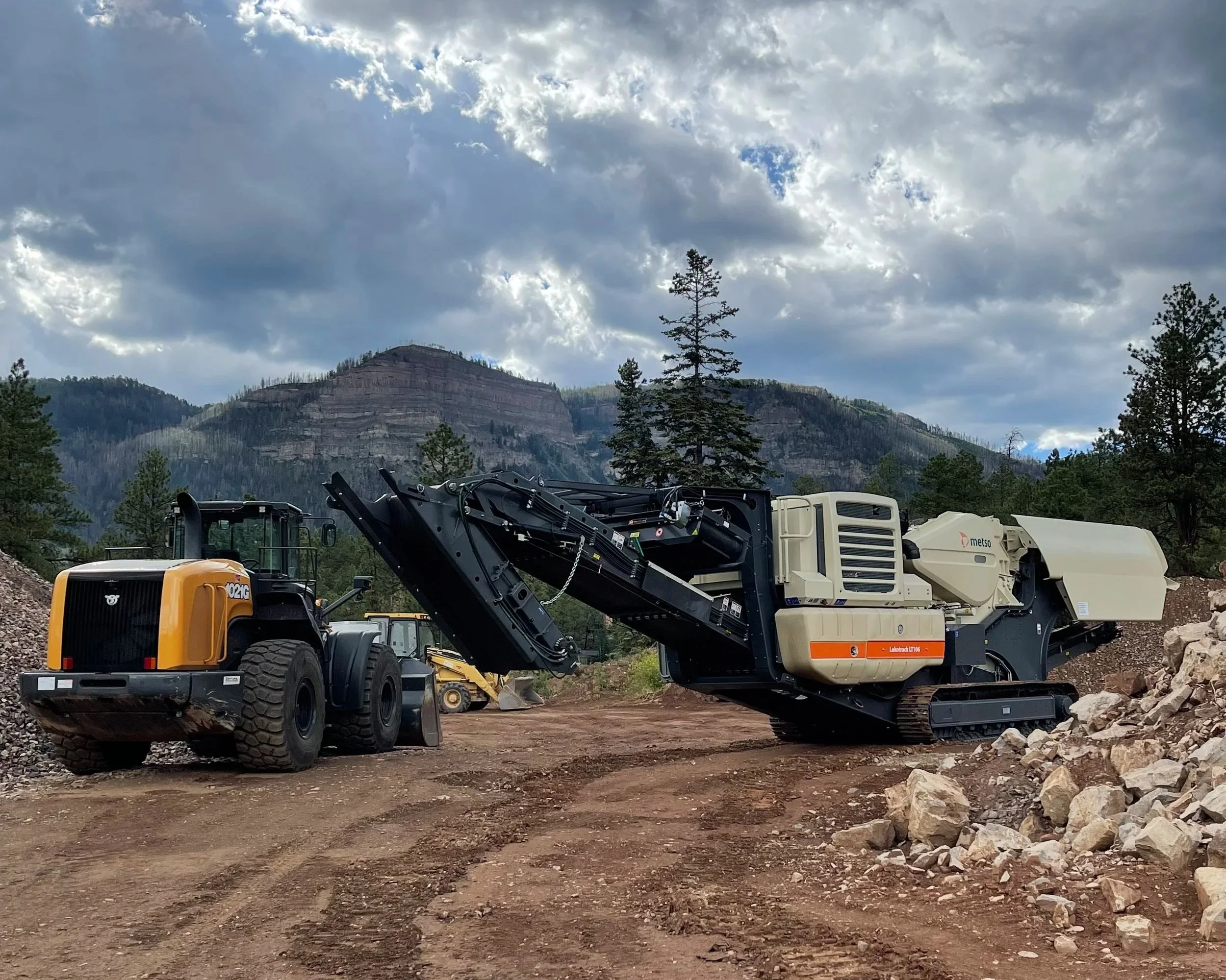 Construction equipment on a dirt lot with rocky terrain, mountains, pine trees, and cloudy sky in the background.