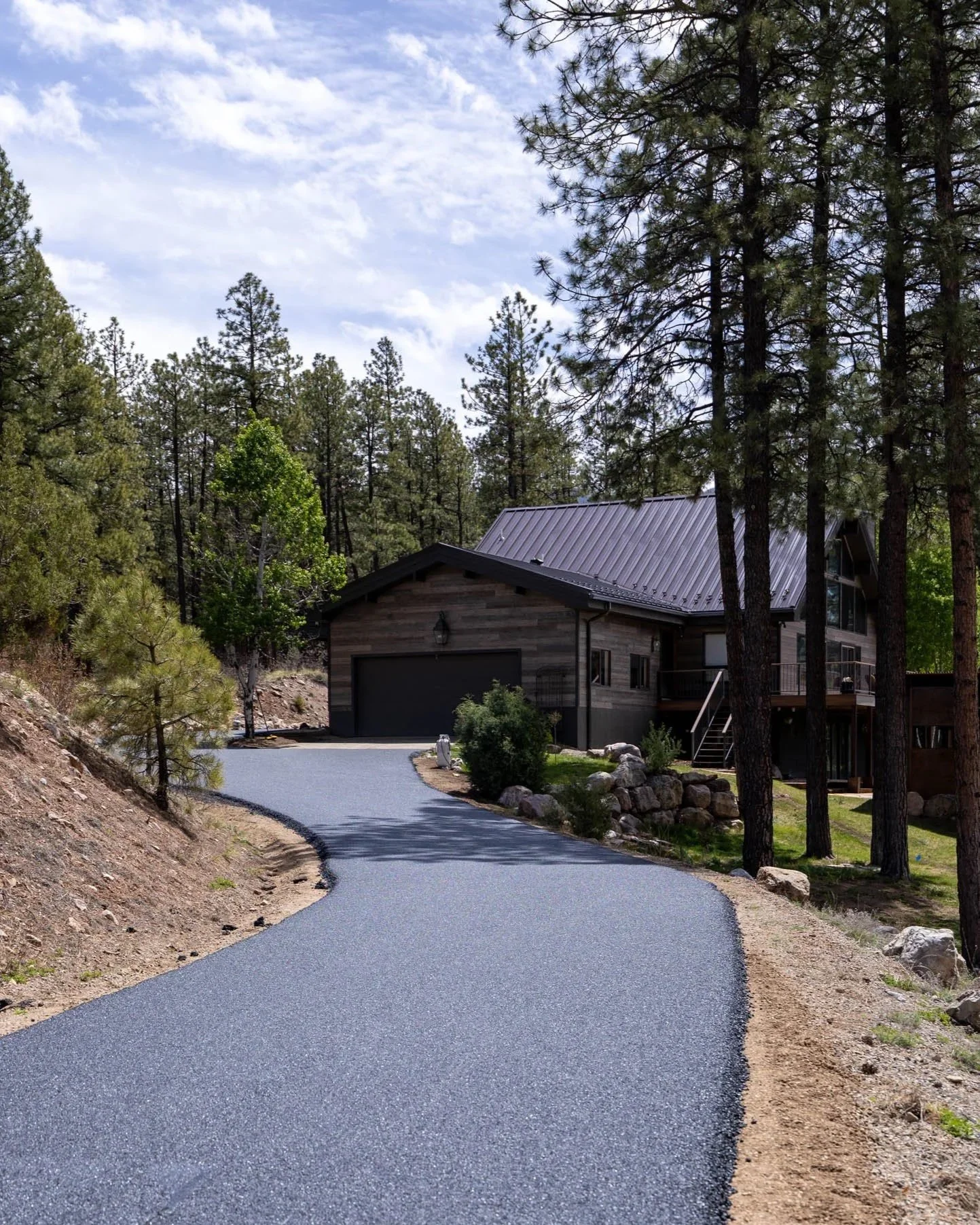 A winding asphalt driveway leading to a wooden house with a metal roof, surrounded by tall pine trees and a partly cloudy sky.