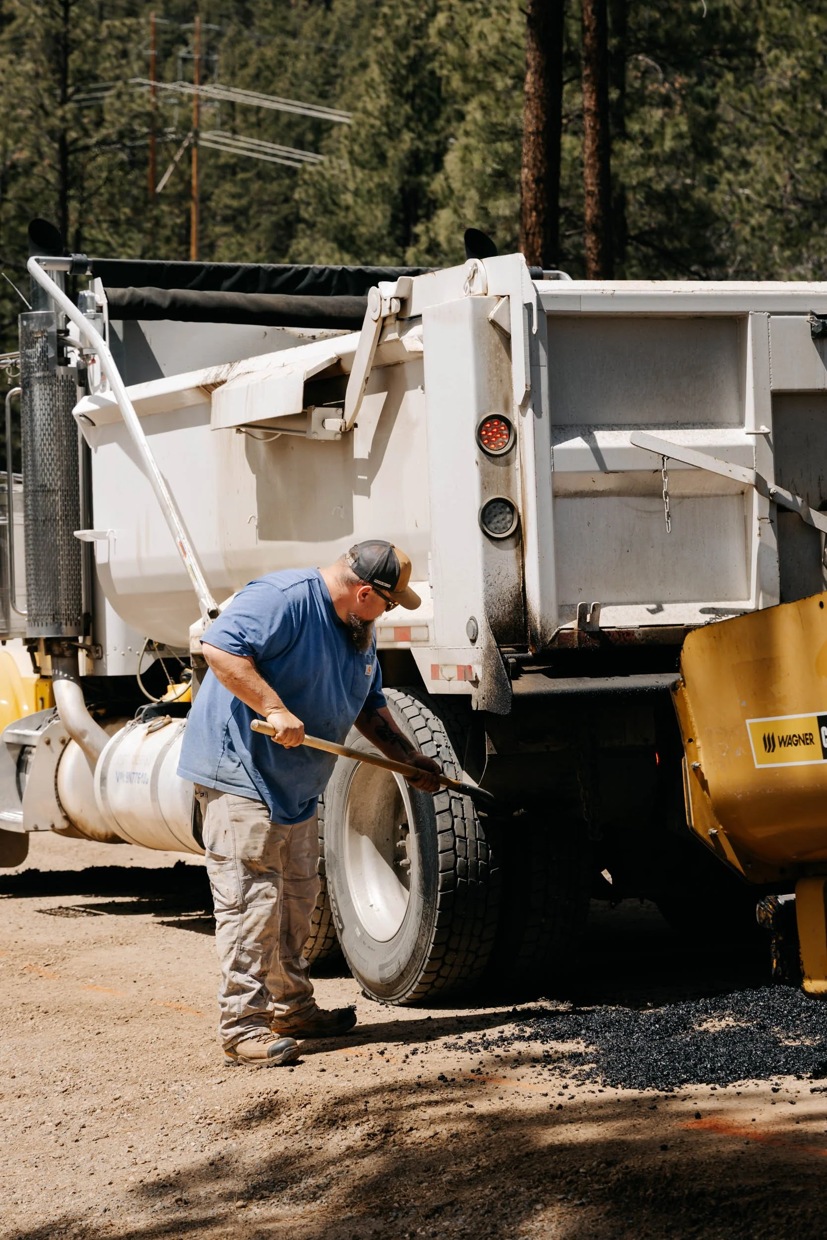 A worker in a blue shirt and beige pants is working on a large white construction truck, possibly repairing the tire or inspecting it. The truck is on a dirt ground with trees and power lines in the background.