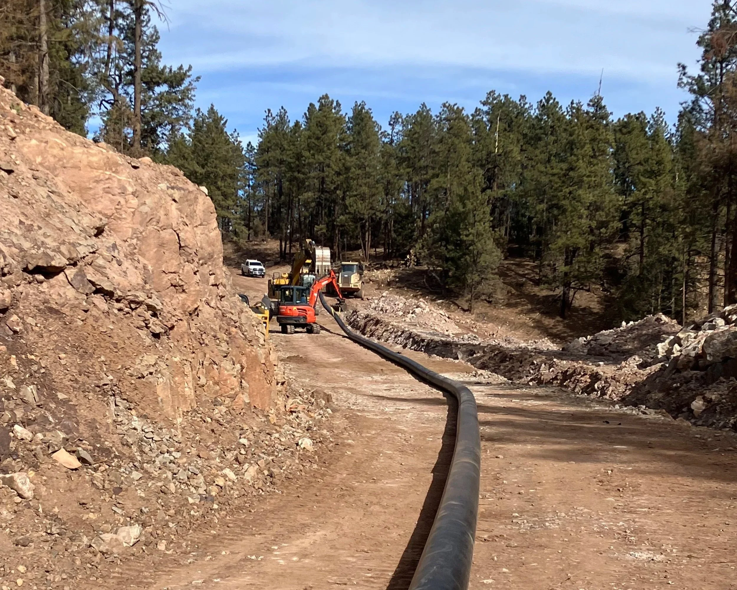 Construction site with large pipe laid on dirt ground, construction equipment, and a forest of tall pine trees in the background.