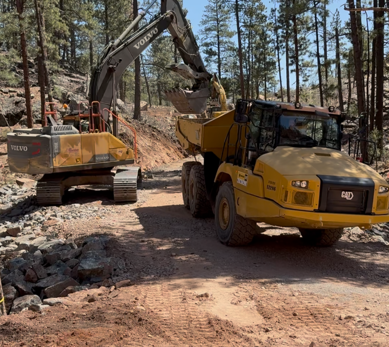 A construction site in a forest with a yellow Caterpillar dump truck loading dirt into a Volvo excavator.