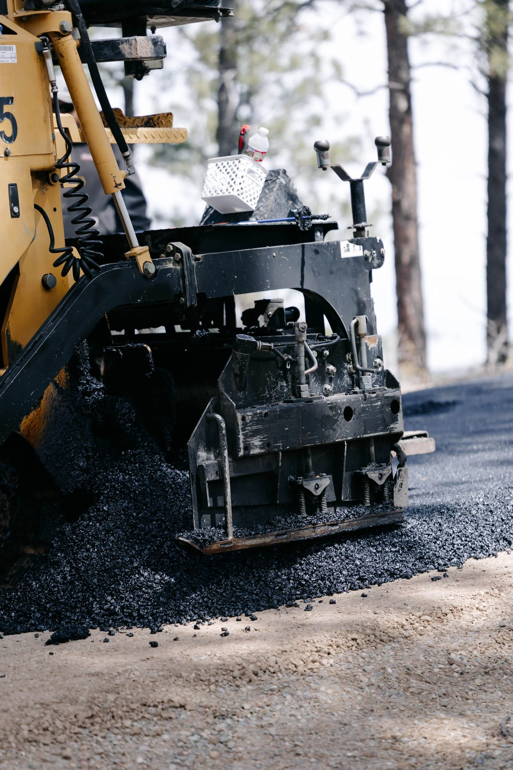 A bulldozer spreading fresh black asphalt onto a road in a forested area with trees in the background.