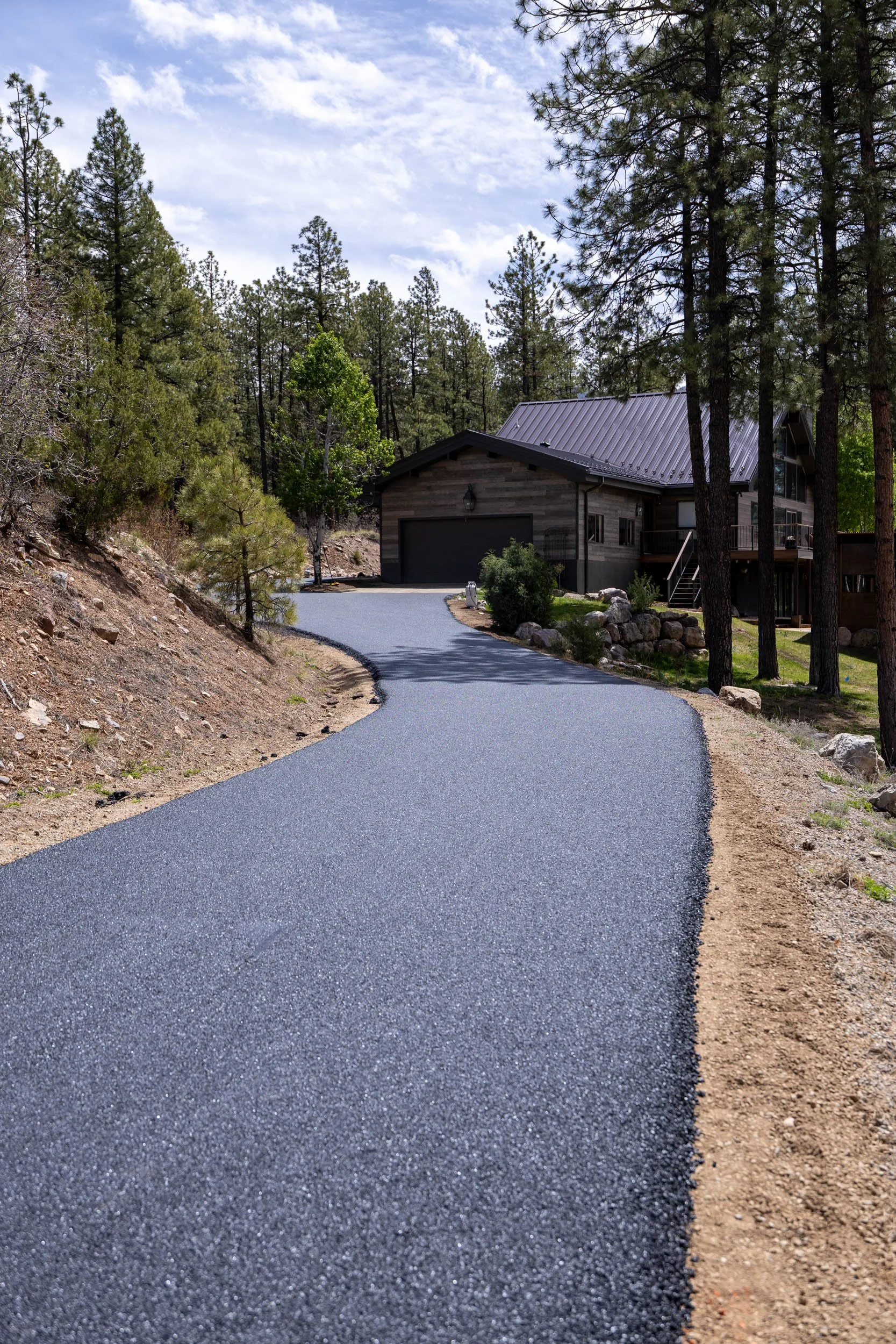 A winding paved driveway leads to a modern house with a metal roof, surrounded by tall pine trees and rocky terrain in a forested area under a partly cloudy sky.