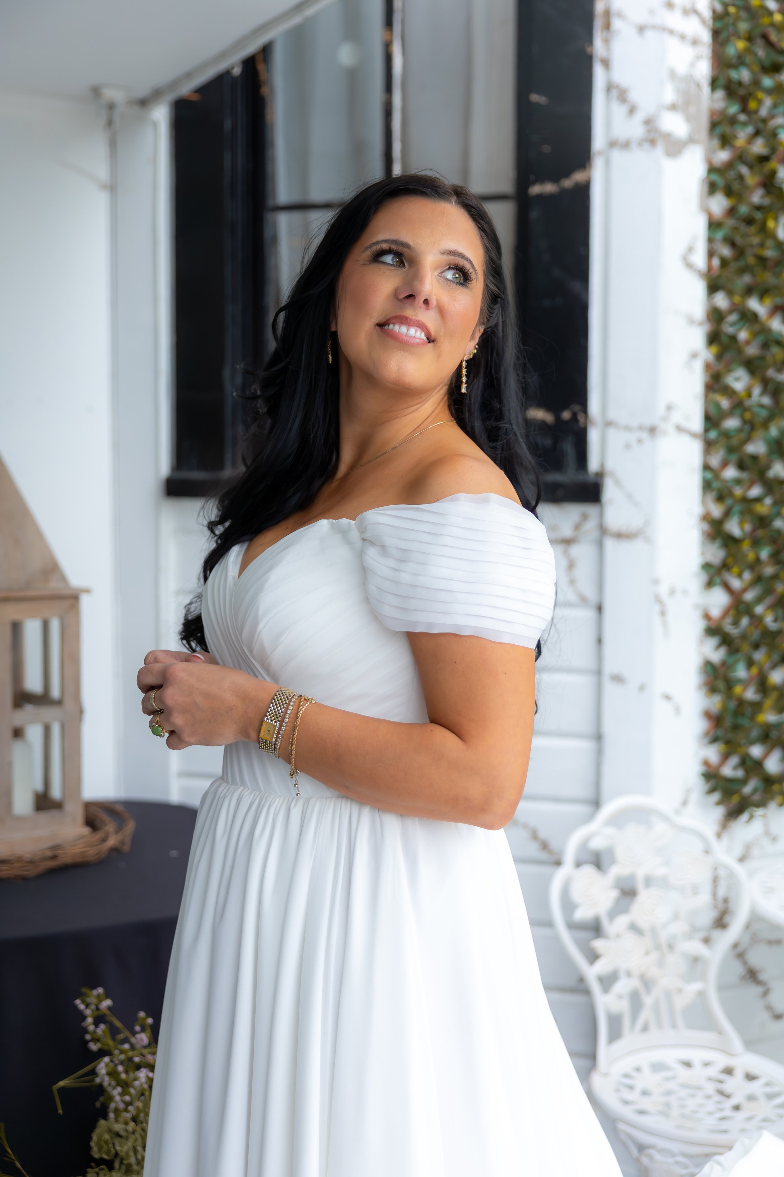 A woman with long dark hair wearing a white dress with puffed sleeves, standing outdoors with a white and black background and leafy plants, smiling while looking sideways.