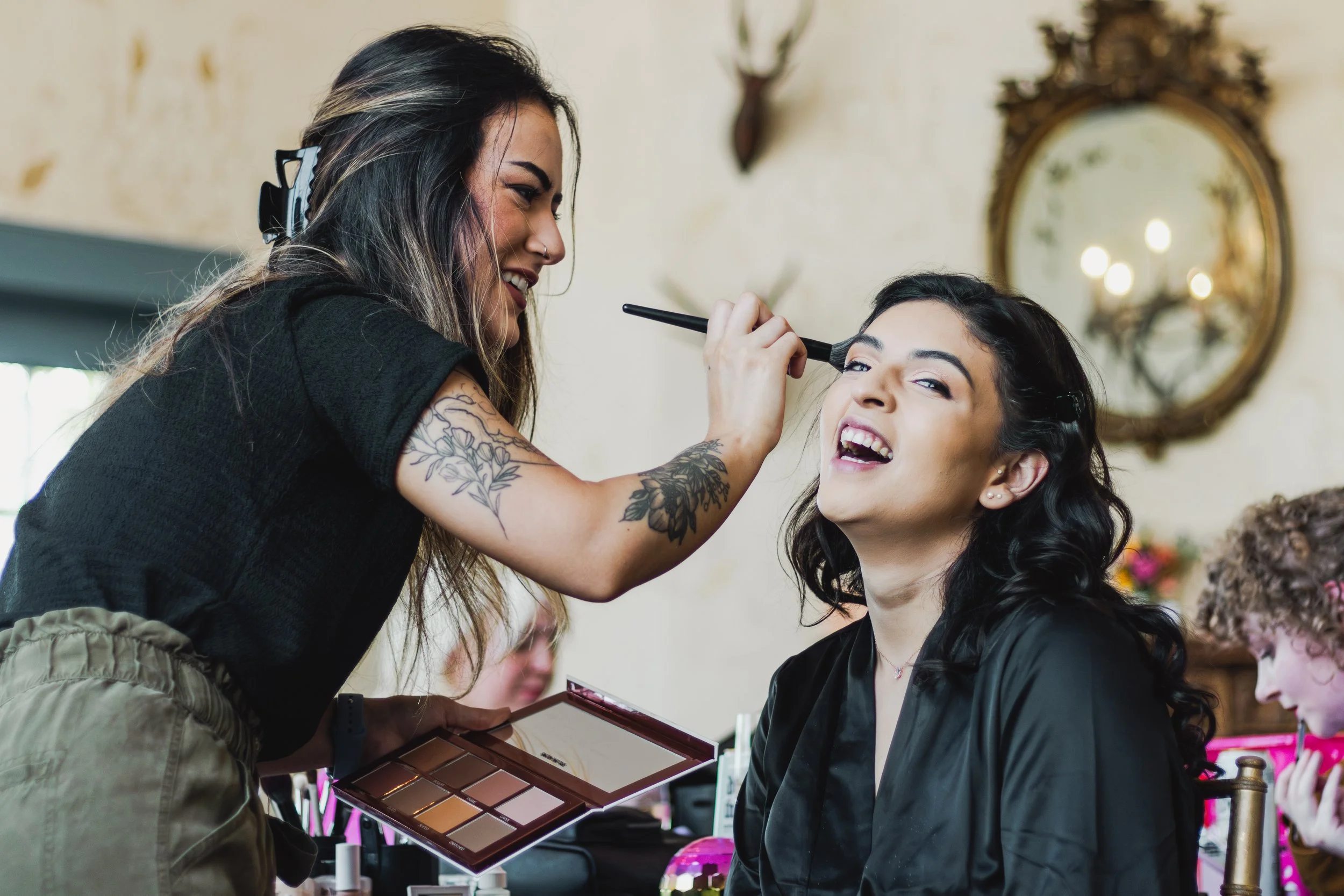 A woman getting her makeup done by a makeup artist, who is smiling and holding a palette of makeup. The woman is smiling as the makeup artist applies eye makeup with a brush.