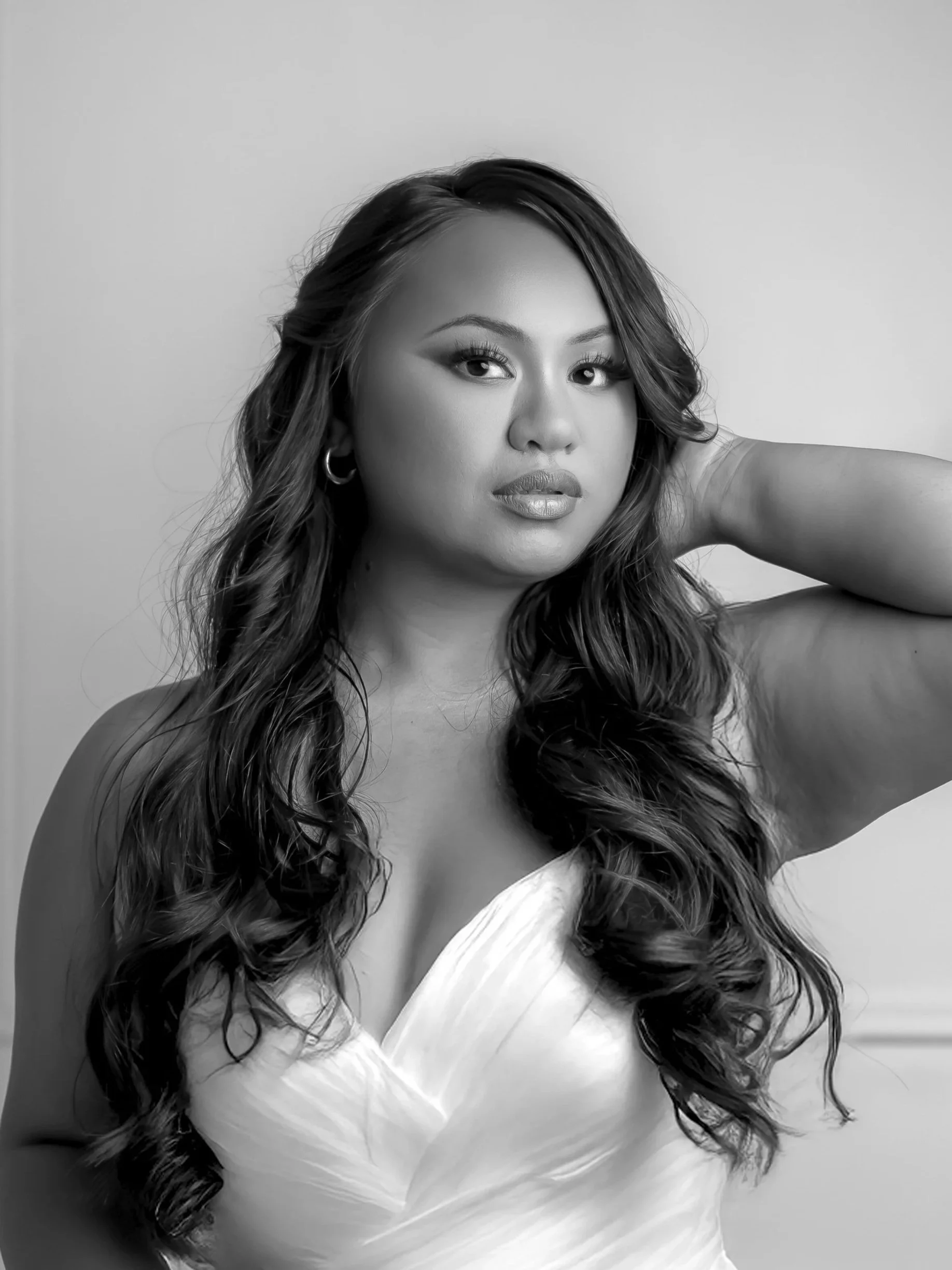 Black and white portrait of a woman with long wavy hair, wearing a strapless dress and hoop earrings, posing with one arm raised behind her head.
