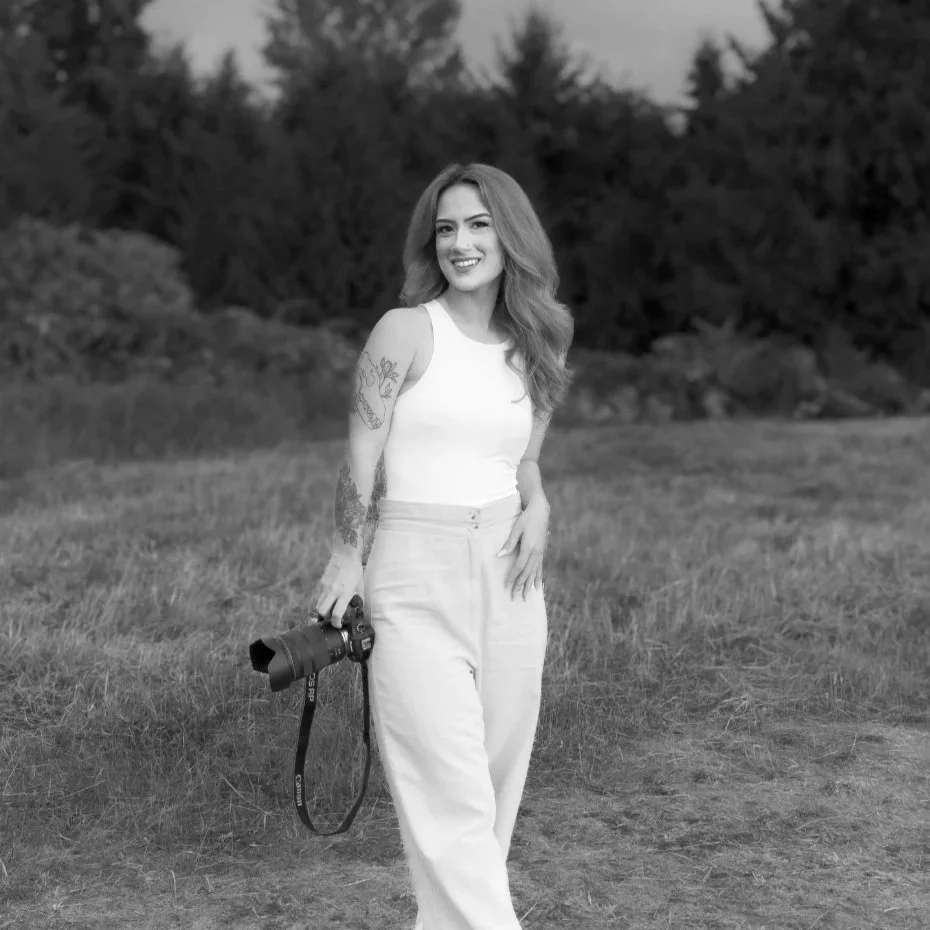 A woman with wavy hair, smiling, stands outdoors in a grassy area, holding a camera in her right hand, with trees in the background.
