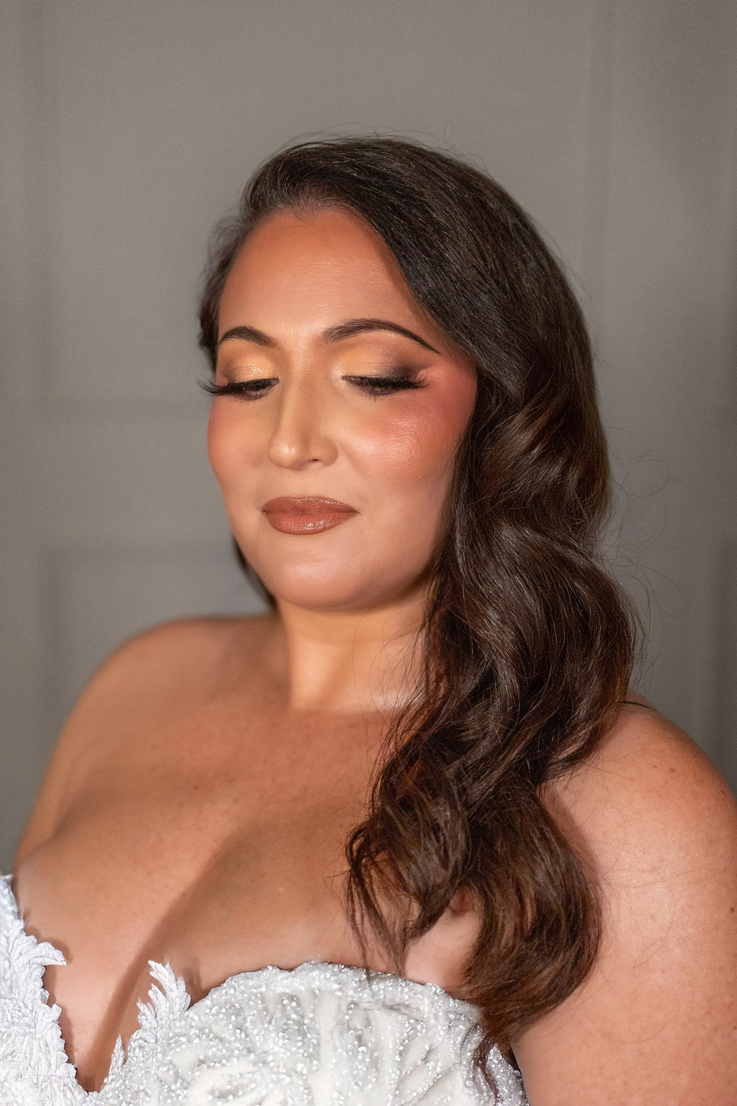 A woman with long, wavy brown hair and makeup, wearing a white, pearl-embellished dress with a deep neckline.