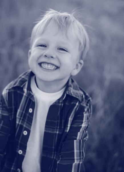 A young boy with blond hair smiling widely, wearing a plaid shirt over a white tee, outdoors.