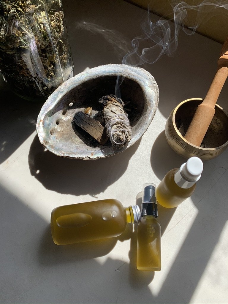 A collection of objects on a table, including burning sage or herbs in a shell, a mortar and pestle, and bottles of essential or massage oils.