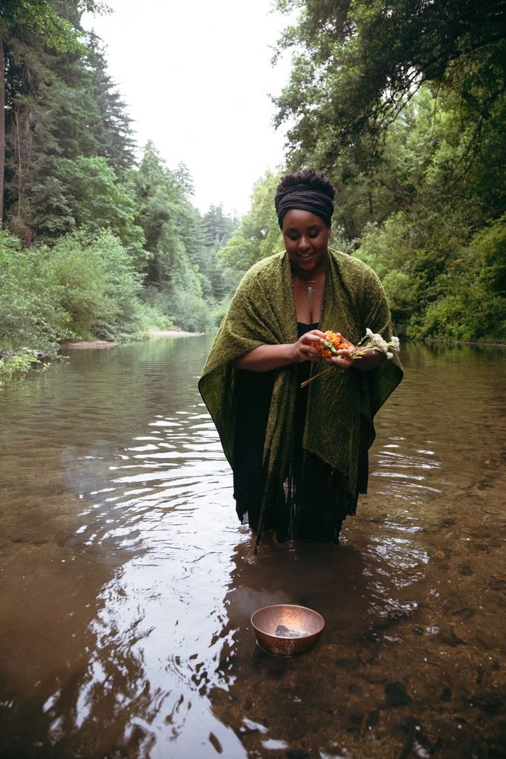 A woman standing in a river surrounded by lush green trees, holding what appears to be a fish and some items, with a small vessel floating in the water nearby.