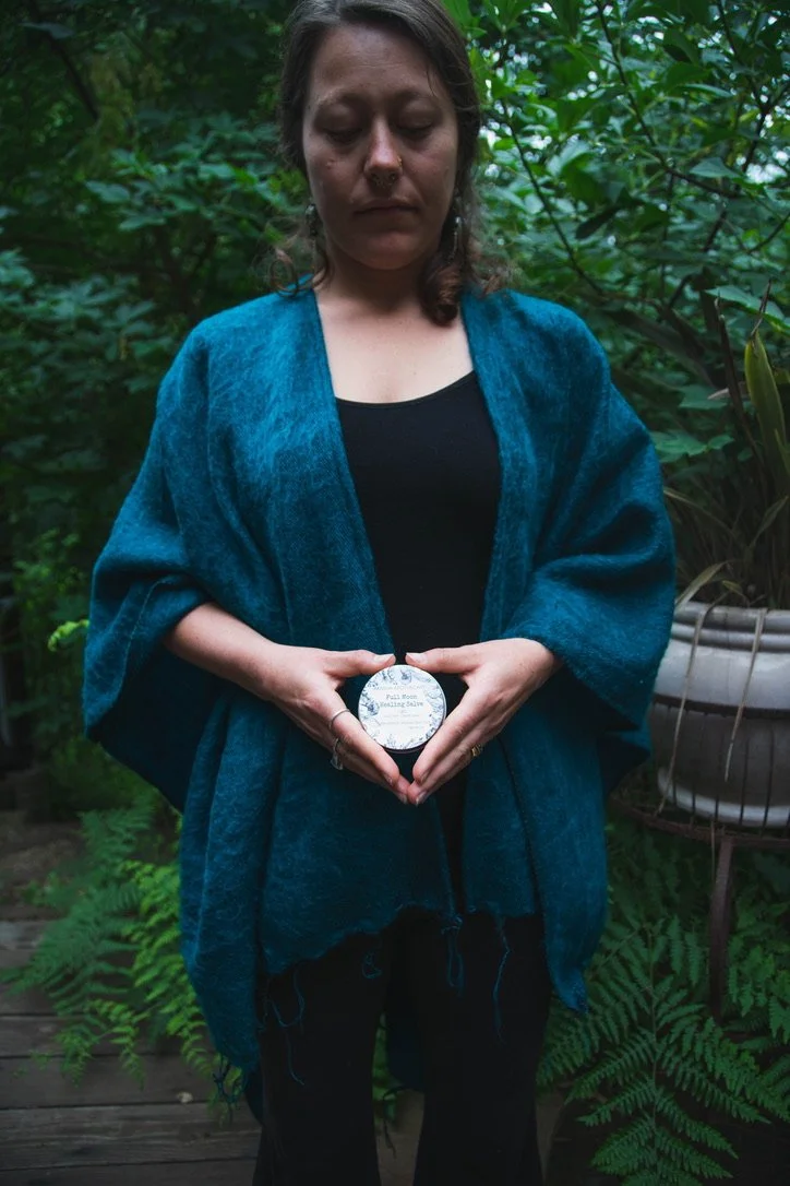 A woman standing outdoors surrounded by green plants, holding a small jar with a label that reads 'Facial Moisturizing Salve'.