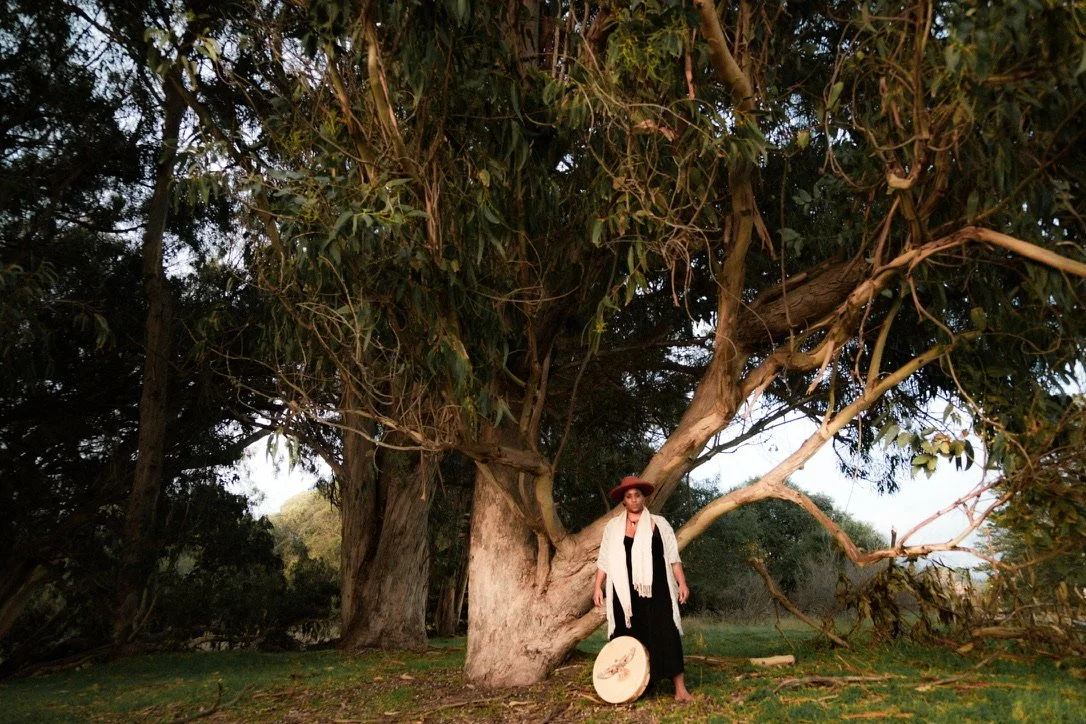 A woman standing under a large tree with sprawling branches, holding a round drum and wearing a hat and scarf.