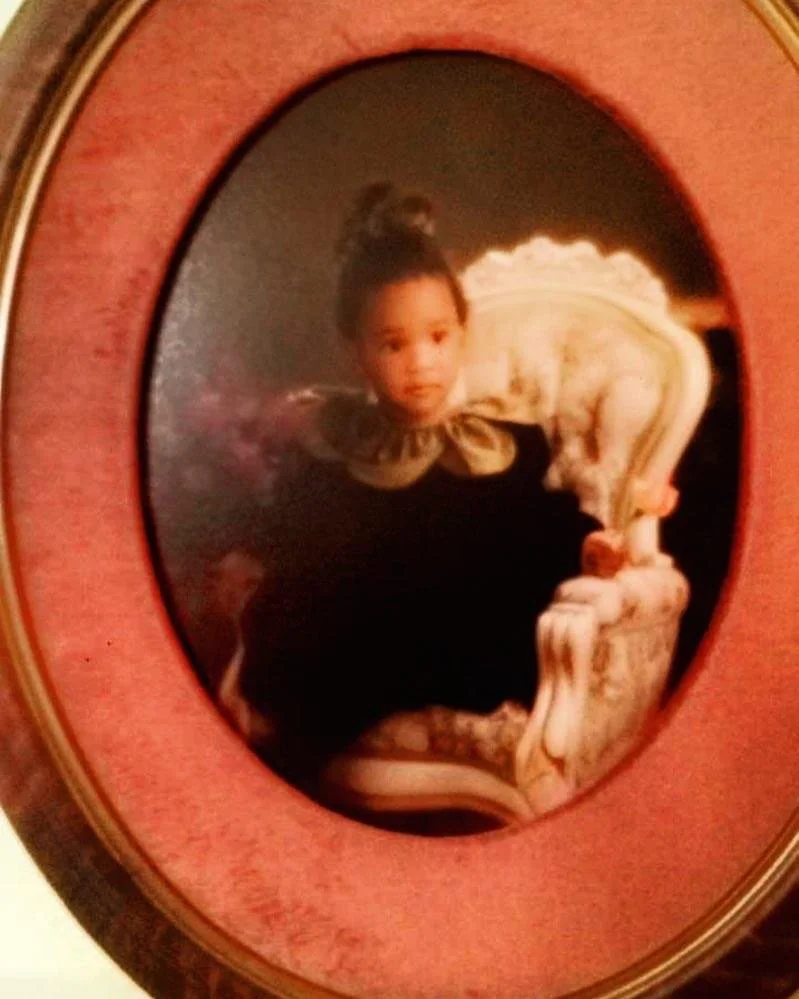 A young girl dressed in a black dress with a bow, sitting on a decorative chair, inside an oval-shaped, pink-framed picture.