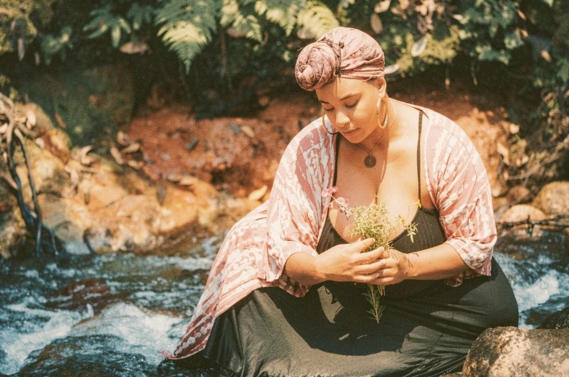 A woman sitting in a stream, holding a plant, with lush green foliage in the background.