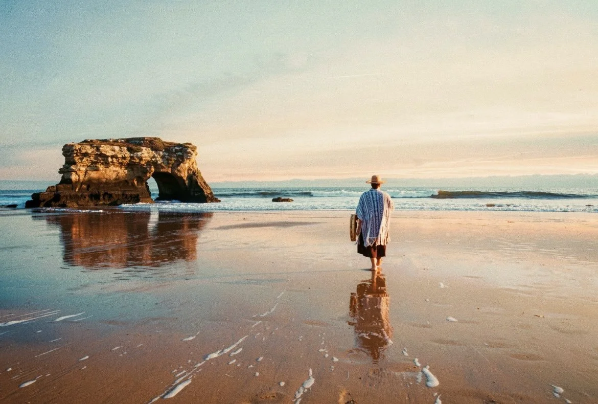 Person walking on the beach toward rock formation at sunset, holding a surfboard, wearing a hat and light-colored cover-up