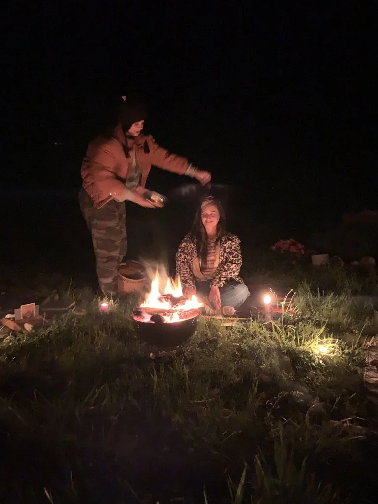 Two women sitting around a small outdoor fire at night, one standing pouring a drink into a cup held by the other woman, with candles and plants nearby.