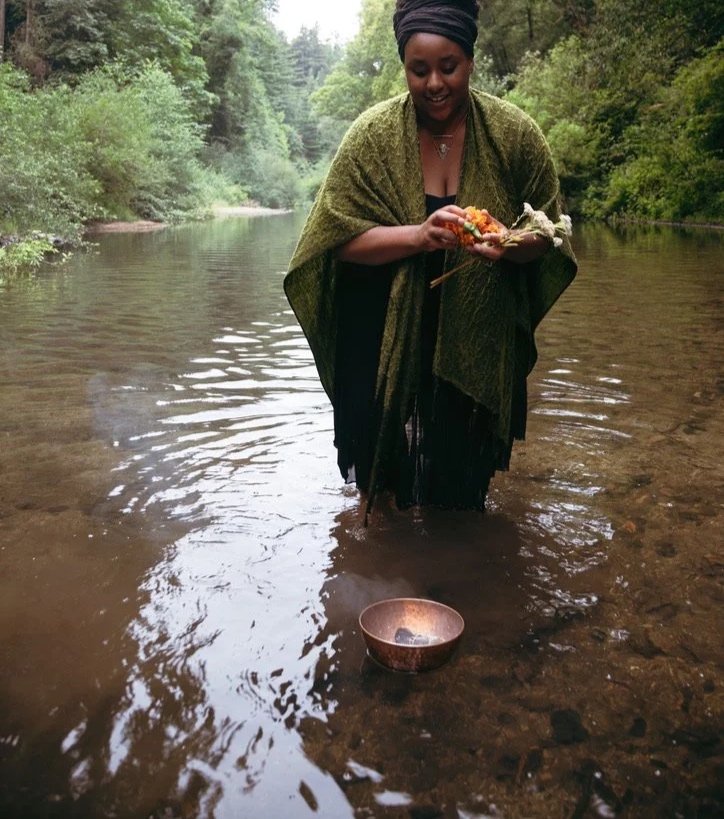 A woman standing in a river with trees in the background, holding a plate of food and a small object, with a copper bowl on the water's surface in front of her.