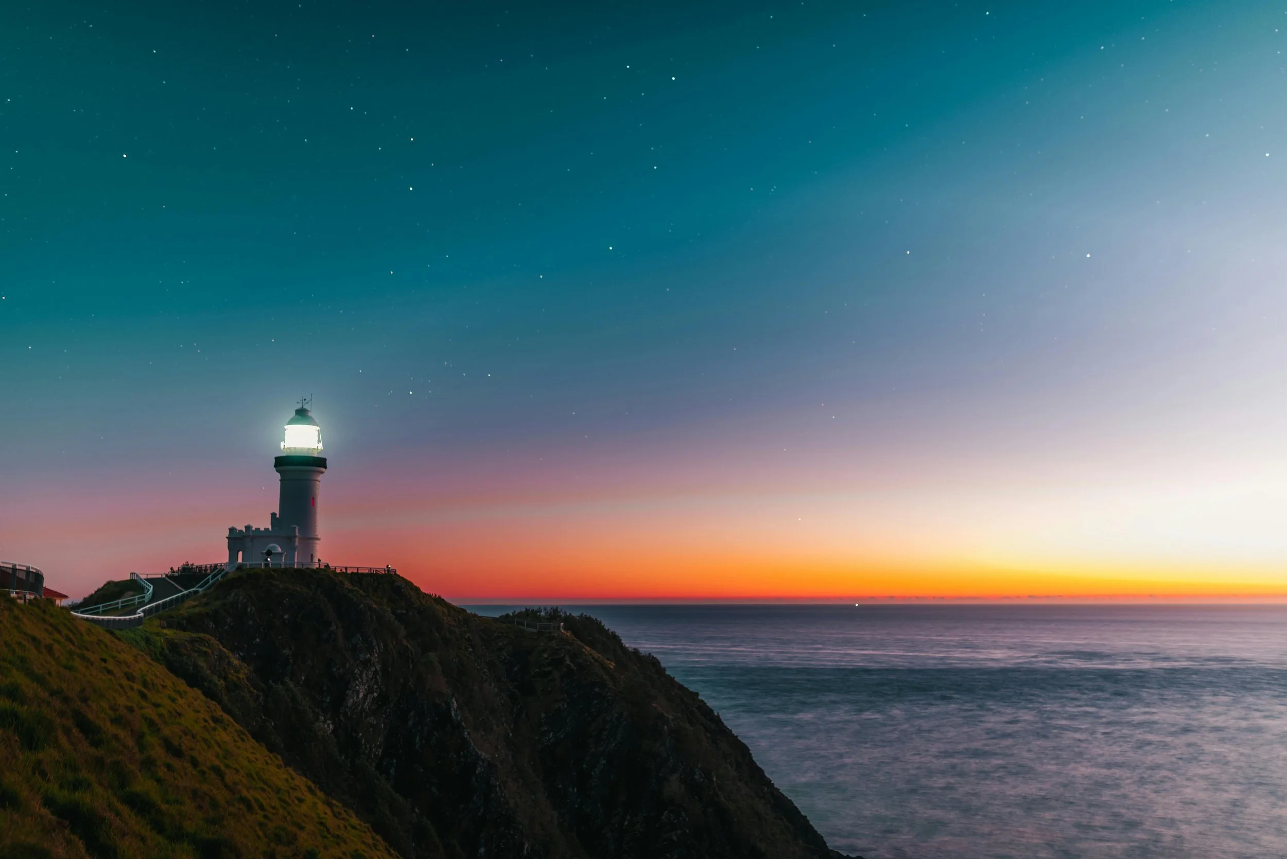 A lighthouse on a cliff at sunset with a colorful sky and stars visible above the ocean.