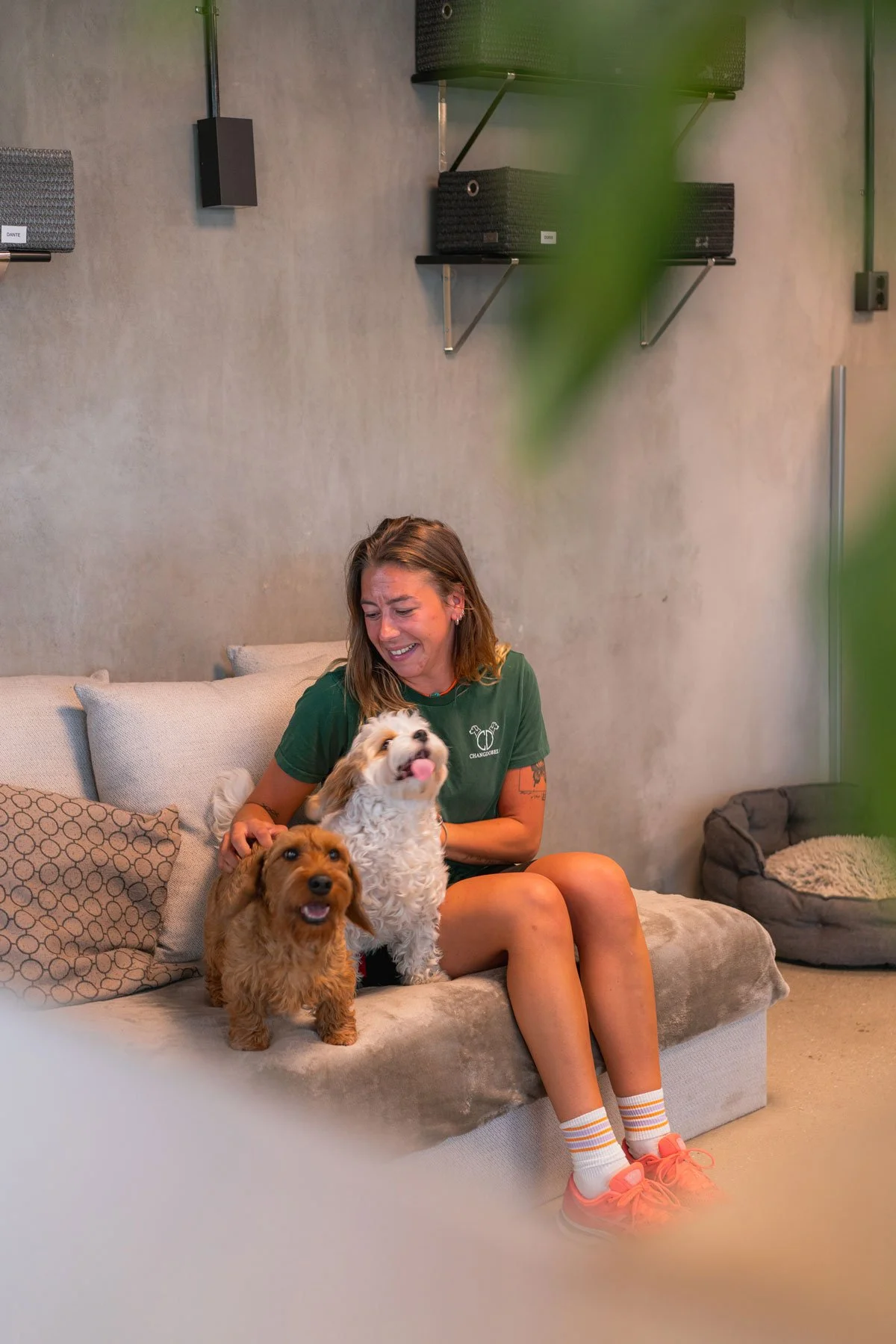 En kvinna sitter på en soffa med tre hundar, två av dem är en liten ljusbrun hund och den andra är en vit hund med bruna fläckar. Kvinnan gråter eller ler med tecken på starka känslor.