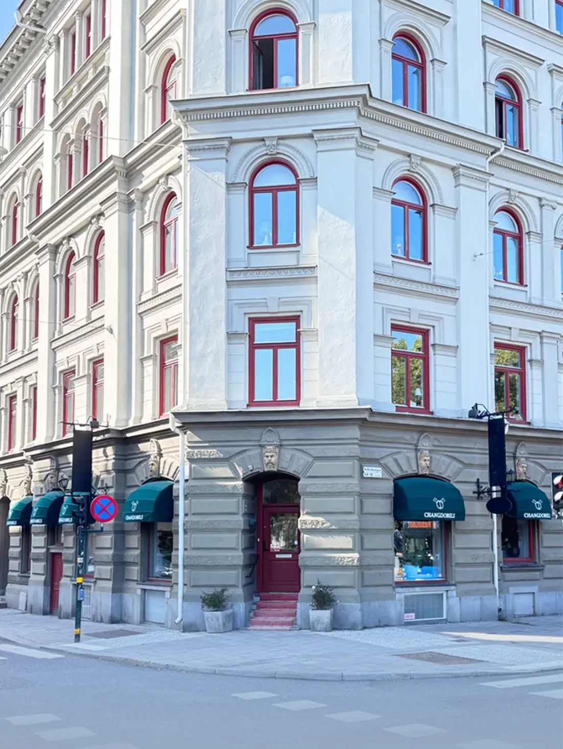 En ljusfärgad, historisk byggnad med moderna skyltfönster och röda fönsterkarmar, idrottsskyltar och en ingång med trappsteg vid hörnet av en gata.