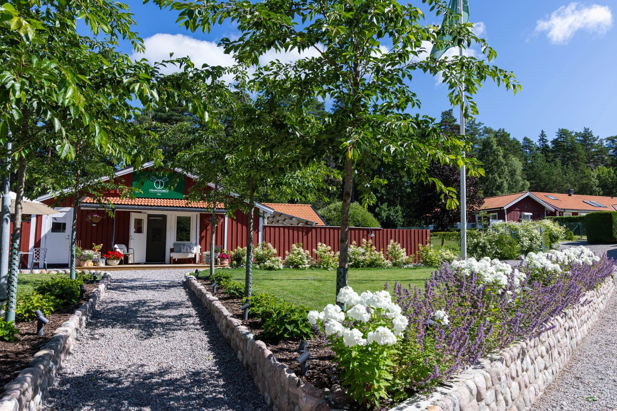 En trädgård med blomsterbäddar fyllda av vita och lila blommor, en grusgång och röda hus med vita fönster och dörrar, omgiven av gröna träd under en klarblå himmel.