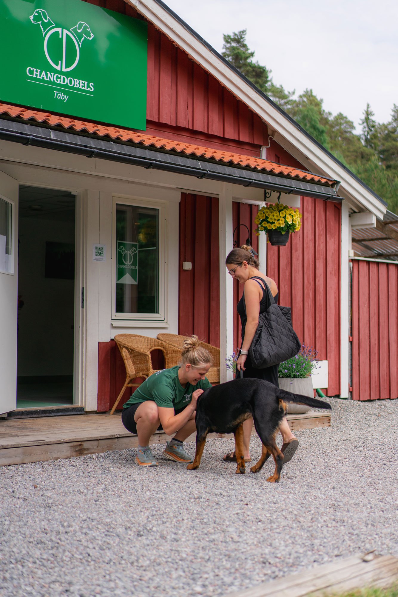 Två kvinnor leker med en hund utanför en röd träbyggnad med blomkrukor och ett grönt tecken på dörren.