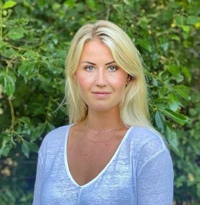 Ung kvinna med blont hår och blå ögon, bär en ljusblå tröja, står framför gröna växter.