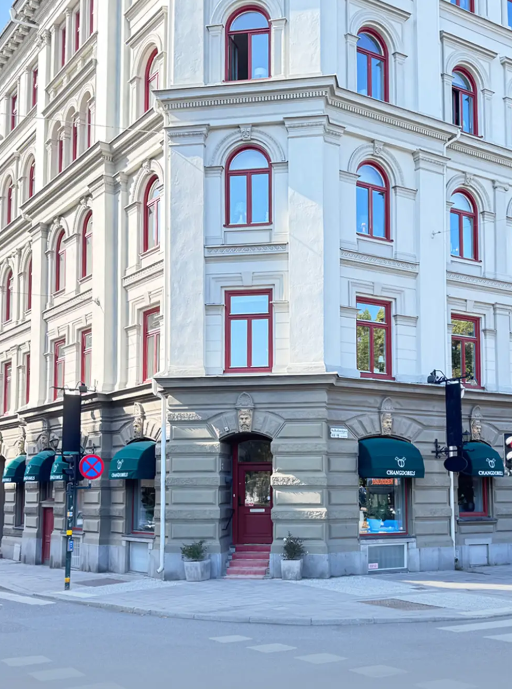 En vit byggnad med många fönster med röda ramar, utsmyckningar och ett ingångsport, samt några gröna växter utanför. Det är en stadsgata med en vägskylt och inga bilar eller människor synliga.