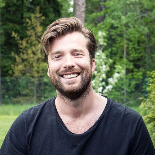 En man med brunt hår och skägg, leende, utomhus med gröna träd i bakgrunden.