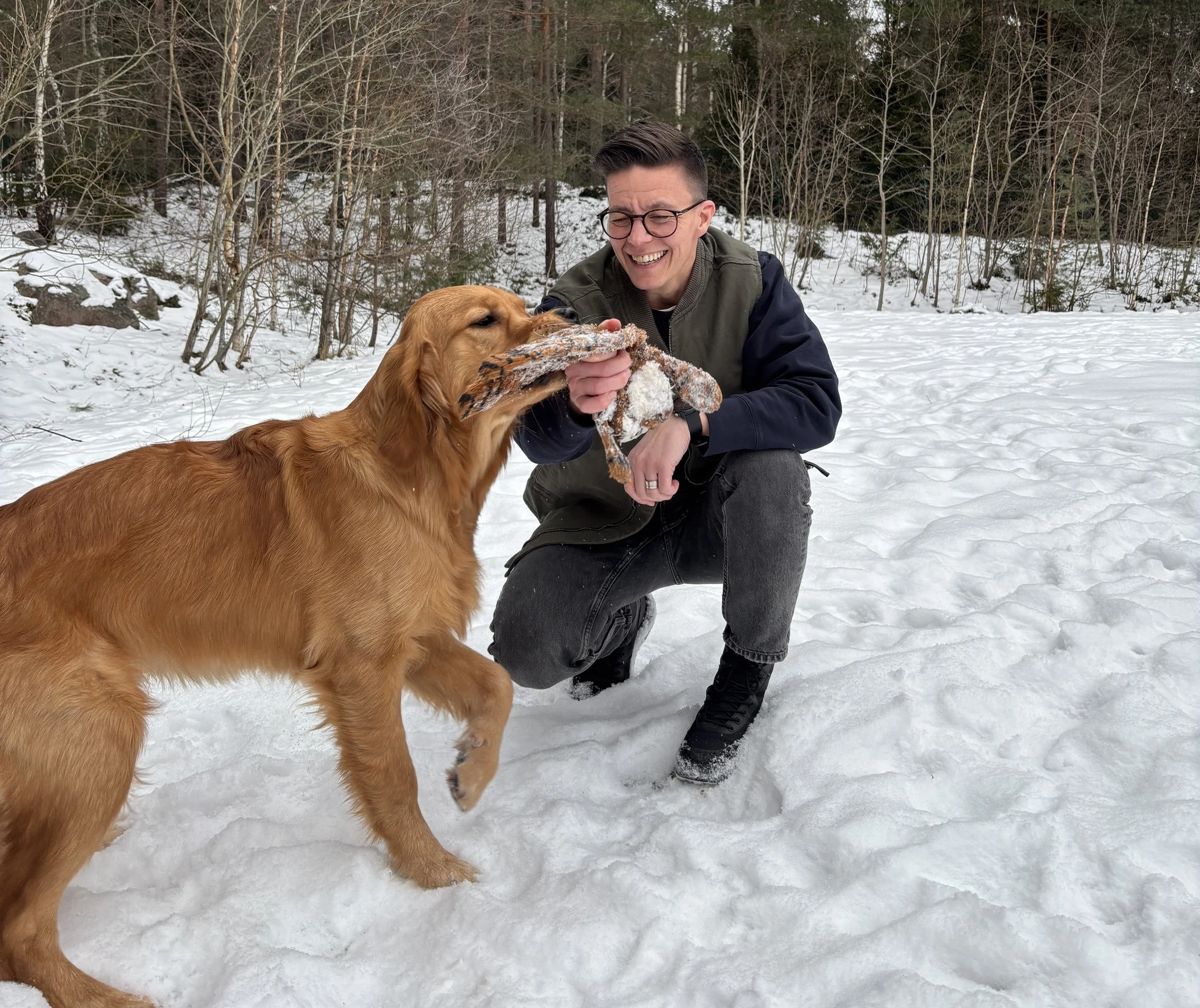 En man med glasögon leker med en golden retriever i snön i en skog.