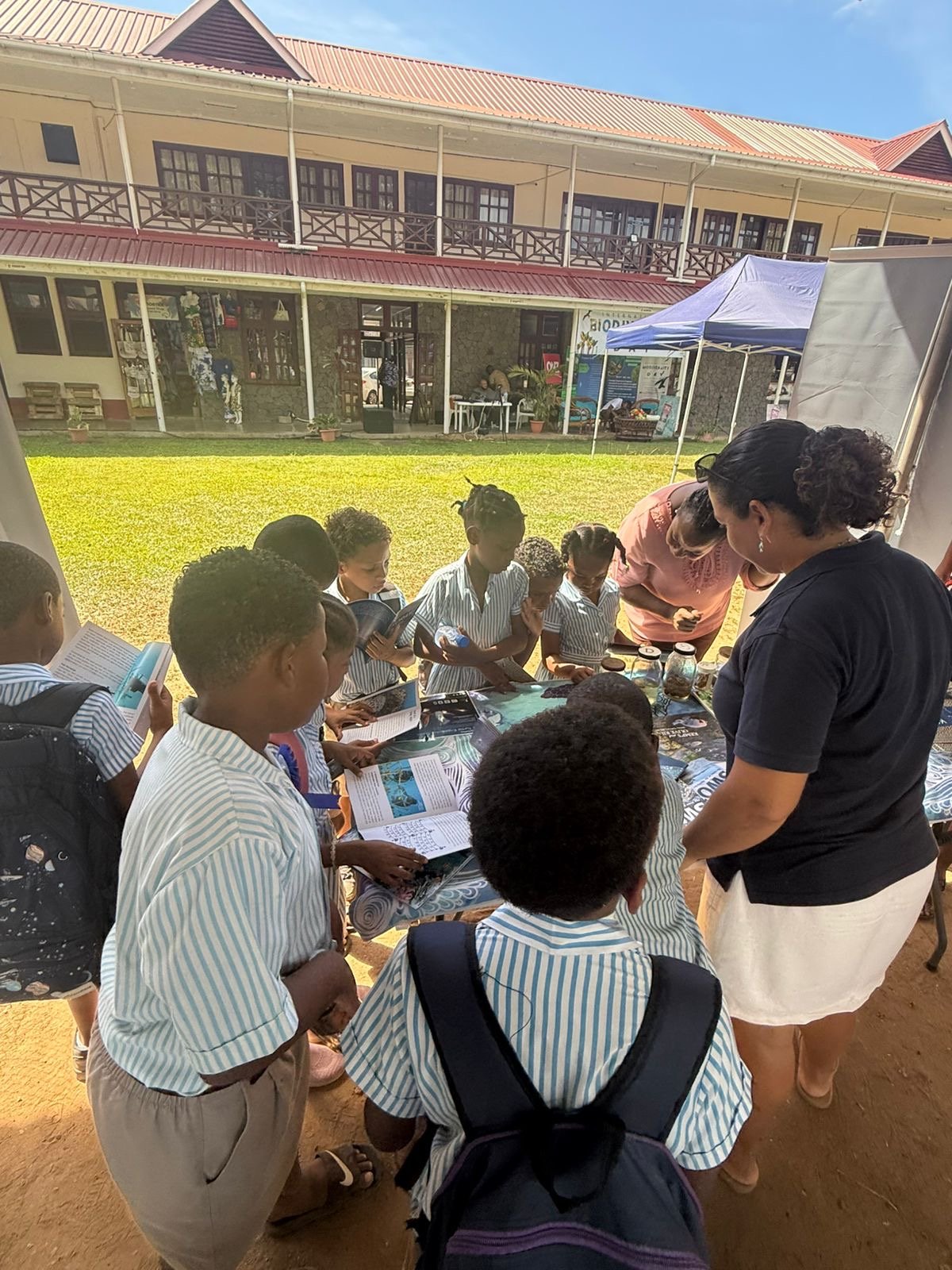 Group of children in school uniforms gathered around a table with books and materials at an outdoor event, with teachers or adults supervising, on a sunny day in a schoolyard.