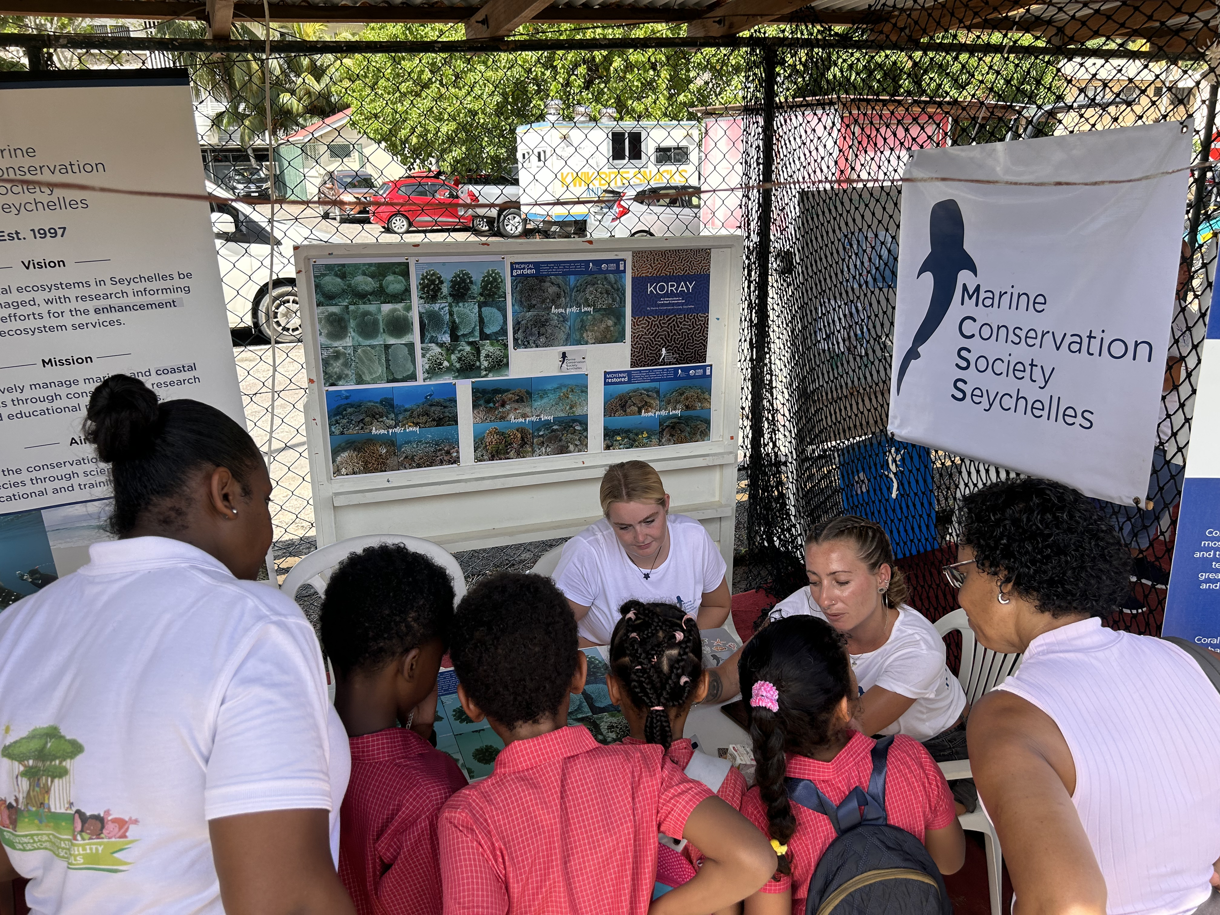 Group of kids and adults gathered around a table at a Marine Conservation Society Seychelles booth, with informational posters and banners about coral reef conservation and marine life in Seychelles.