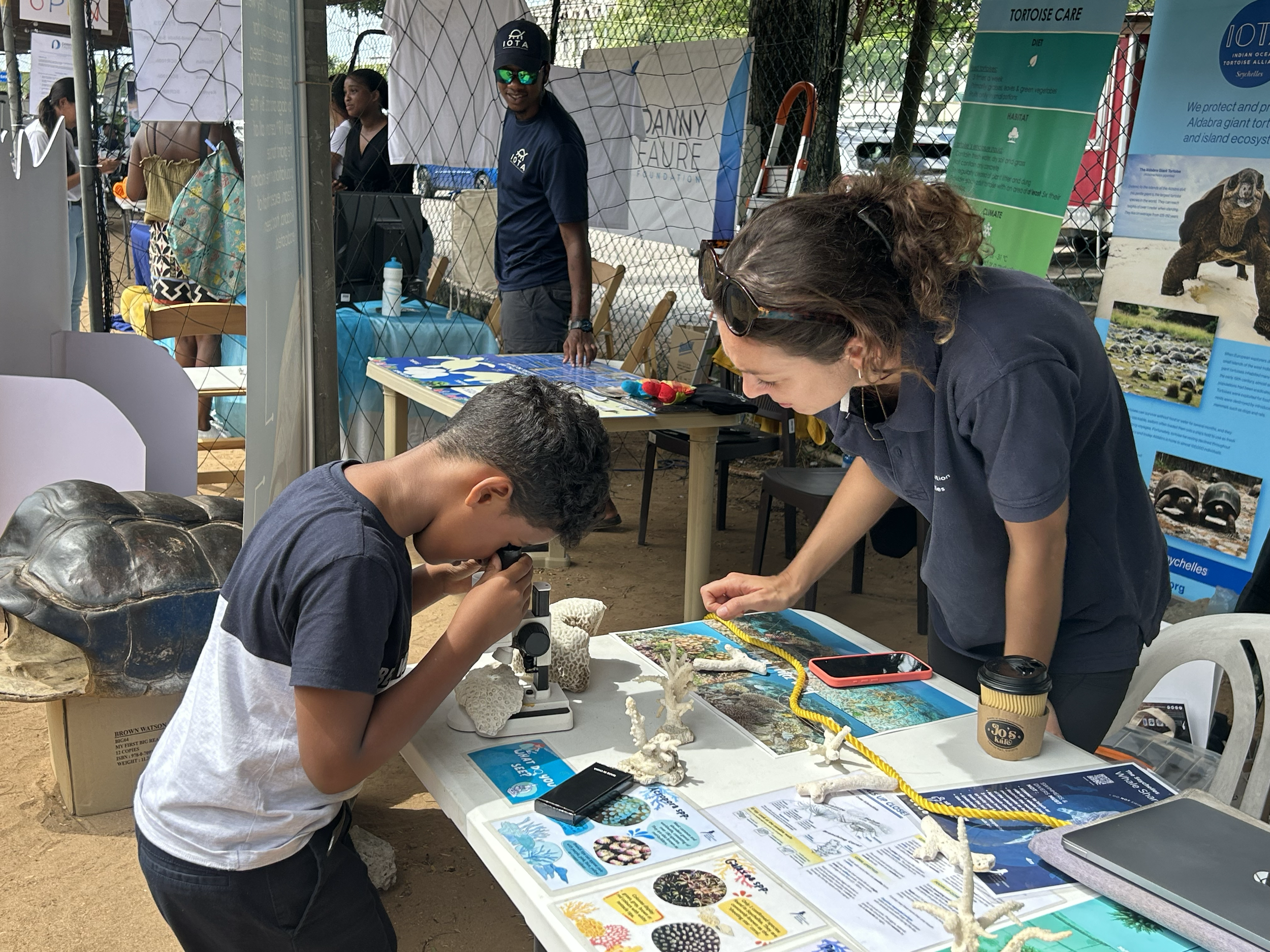 A boy looking through a microscope at an educational display table about marine life, with a woman leaning over to observe. The table has coral samples, brochures, a phone, and a coffee cup, with informational posters about turtles and marine ecosyst