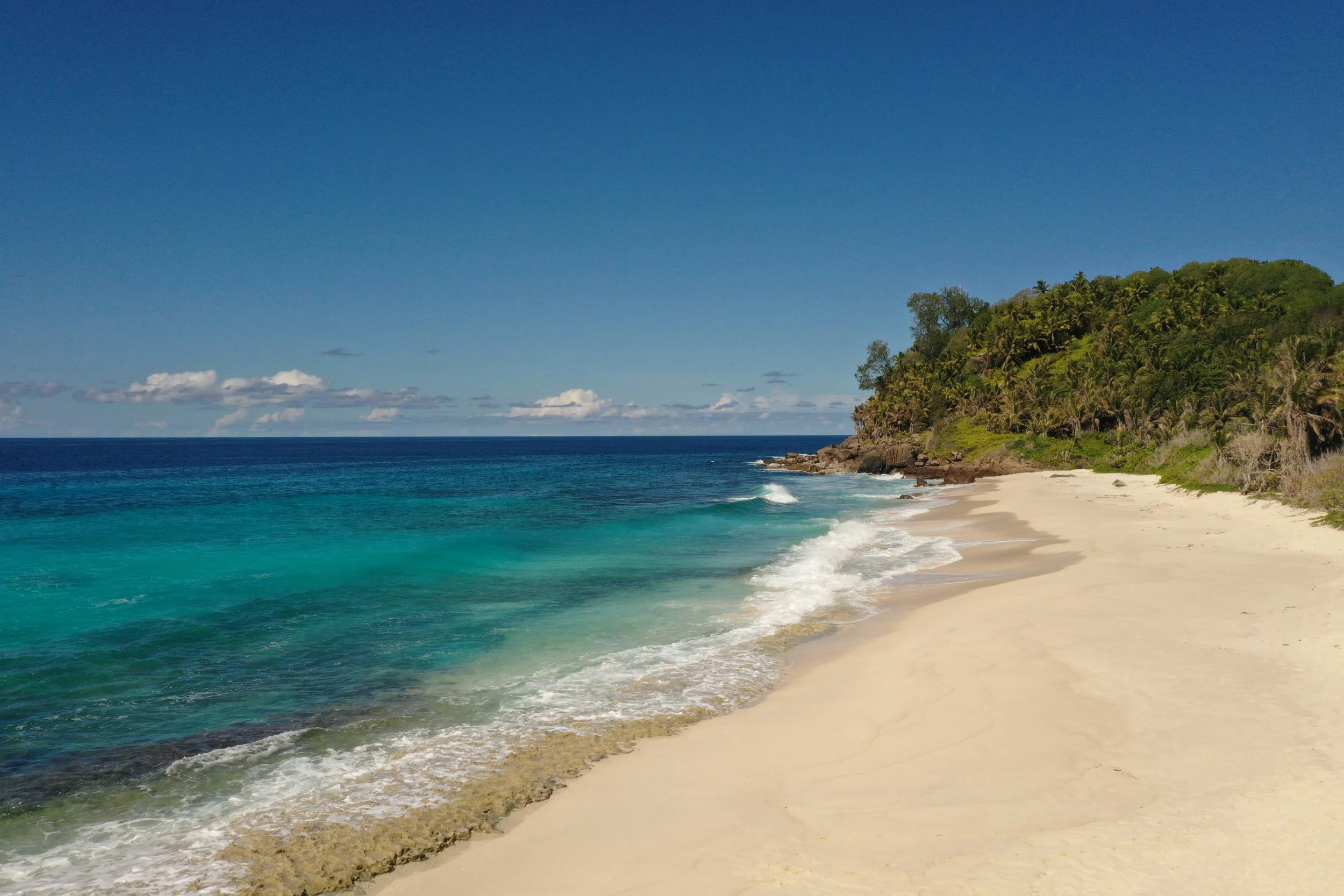 A tropical beach with white sand, turquoise water, and a green hillside in the background under a clear blue sky.