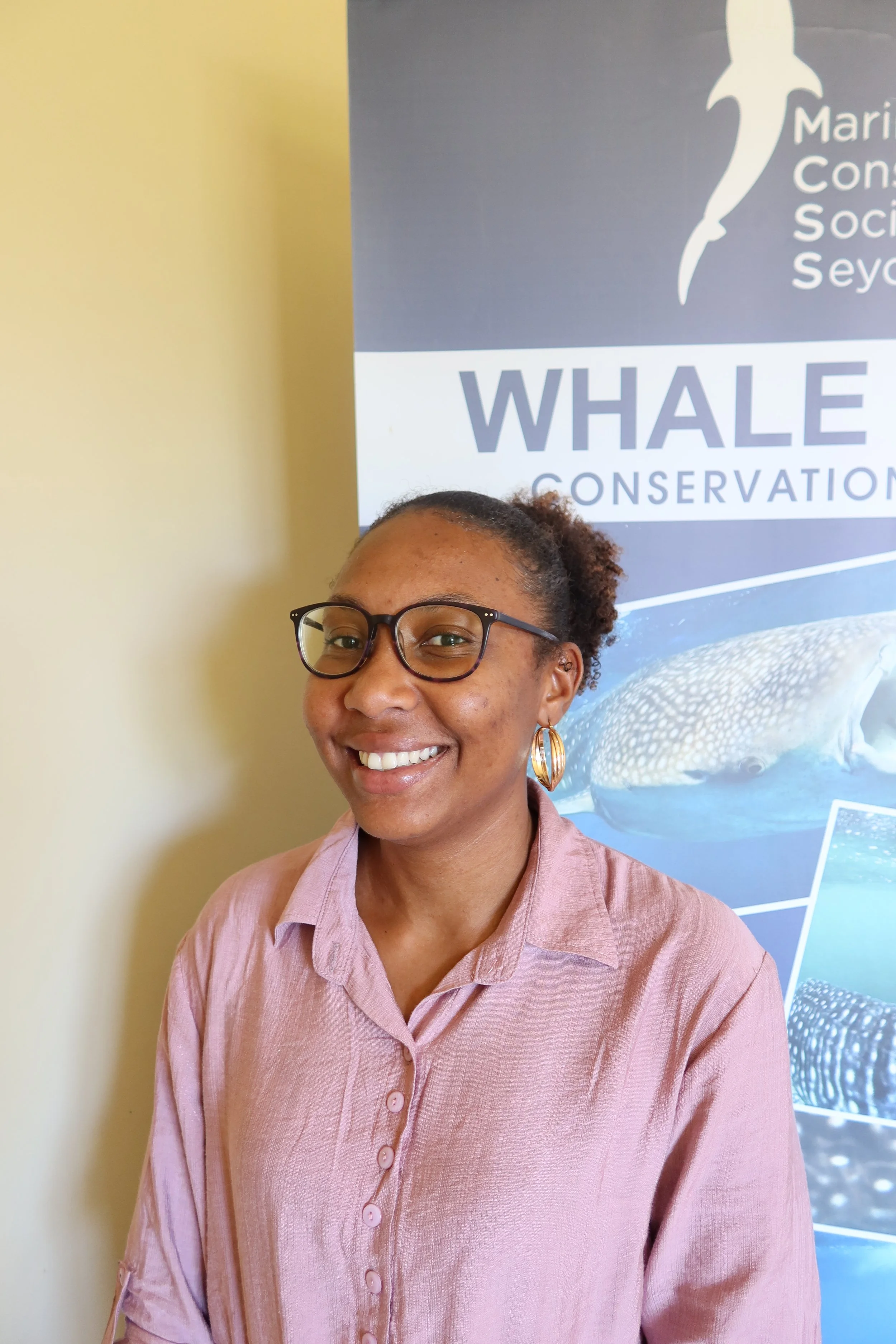 A man with a gray beard and short gray hair, wearing a brown polo shirt and white pants, sitting on a bench against a plain beige wall, with soft natural lighting.