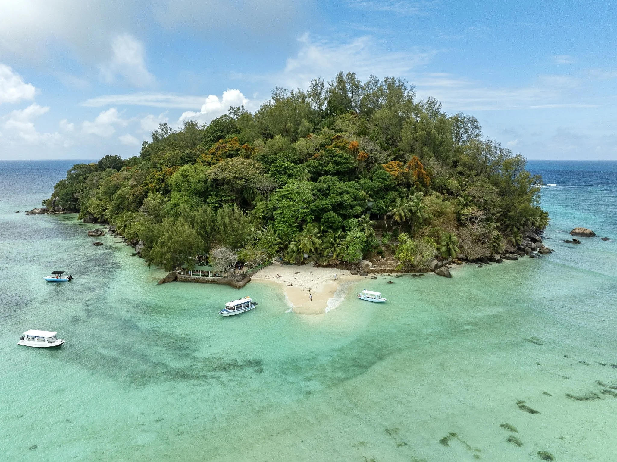 A tropical island with lush green trees, sandy beach, surrounded by turquoise water, with a few boats anchored nearby.