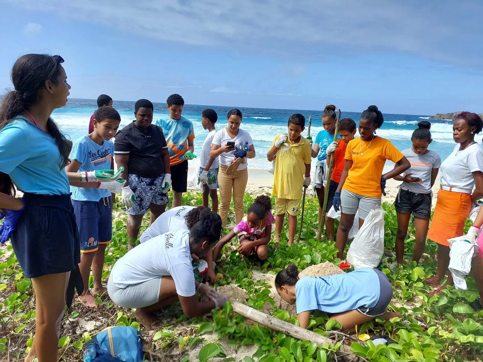 A group of children and young adults on a beach, participating in a planting activity among green bushes, with the ocean and waves in the background.