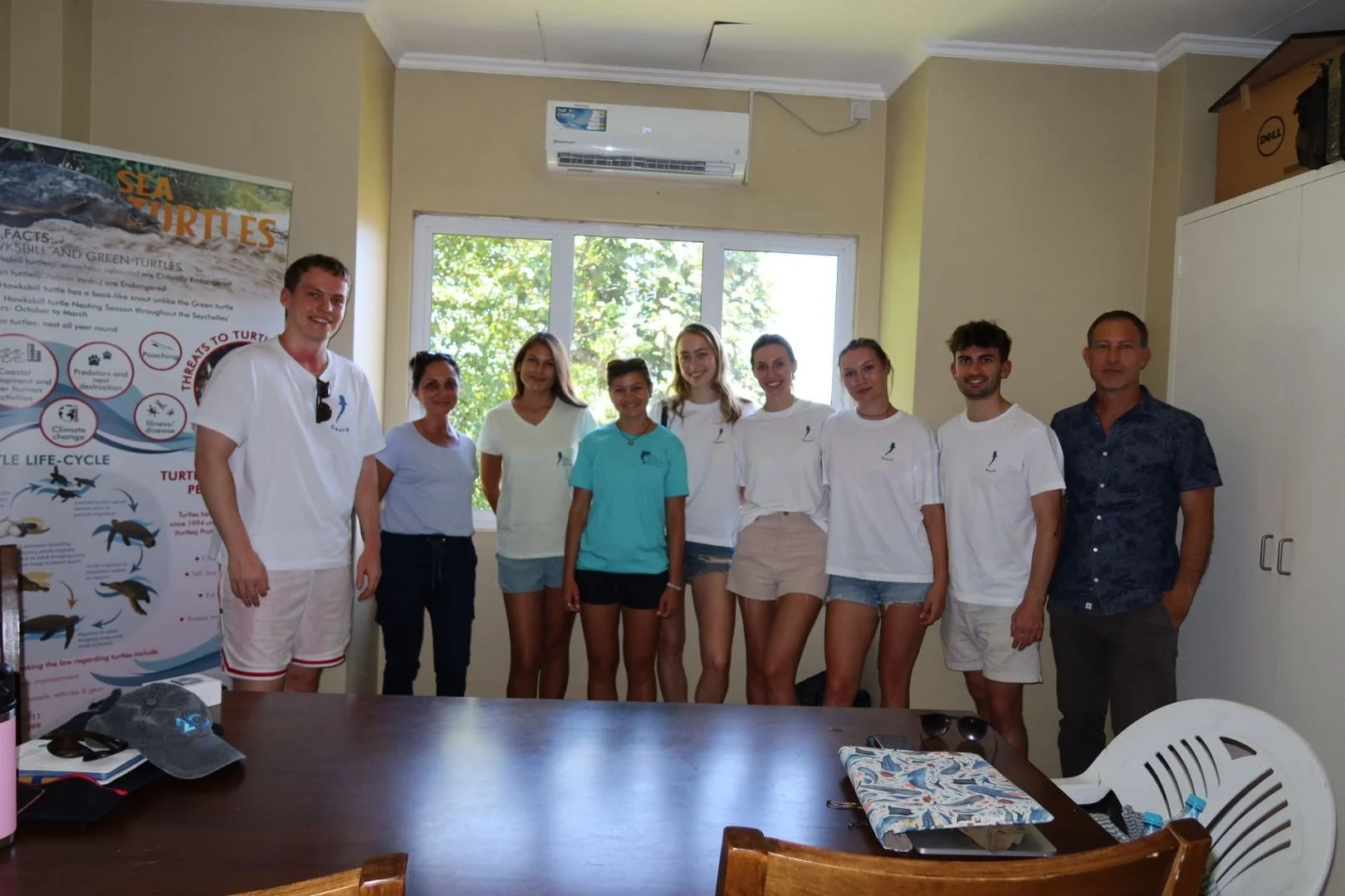 Group of nine people, including six women and three men, standing indoors in front of a window with trees outside. They are smiling and posing for the photo, wearing casual clothing including T-shirts, shorts, and skirts. There is a colorful poster a