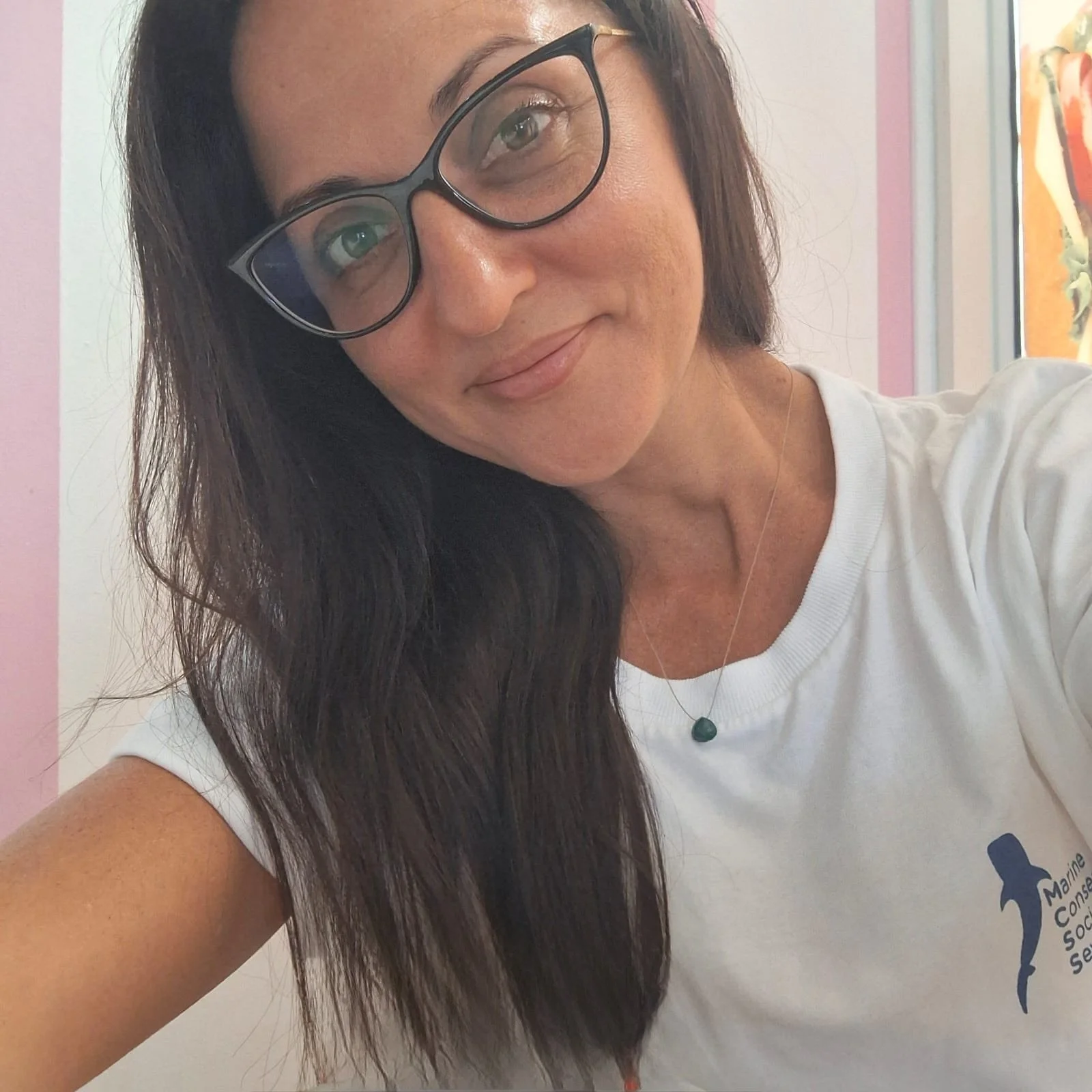 A man with dark, slicked-back hair, wearing a blue knit sweater and beige pants, sitting on a beige cushioned bench against a plain wall, looking to his right with a contemplative expression.