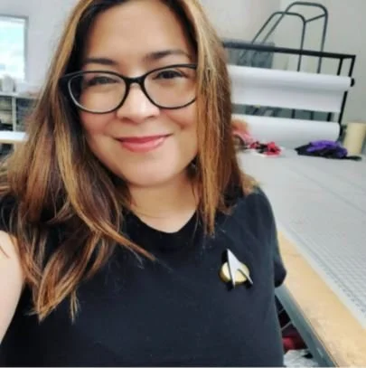 Woman with glasses smiling, wearing a black shirt with a Star Trek insignia, in an indoor setting with tables and materials in the background.