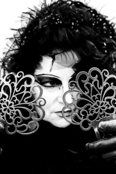A person with dark, spiky hair and makeup holding two decorative circular fans in front of their face, black and white photo